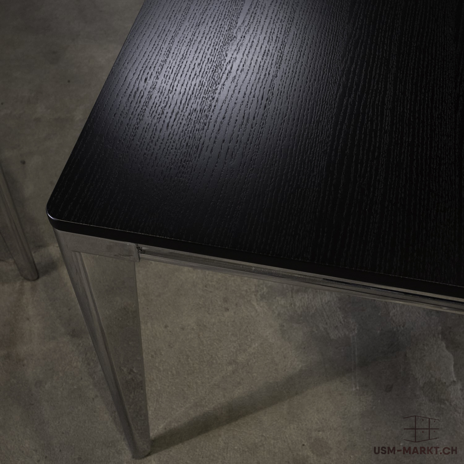 Gros plan sur un plateau de table en bois foncé et veiné, aux angles arrondis. La surface brun-noir laisse apparaître une fine structure du bois. Sous le plateau, on aperçoit un pied de table en métal brillant posé sur un sol en béton gris.