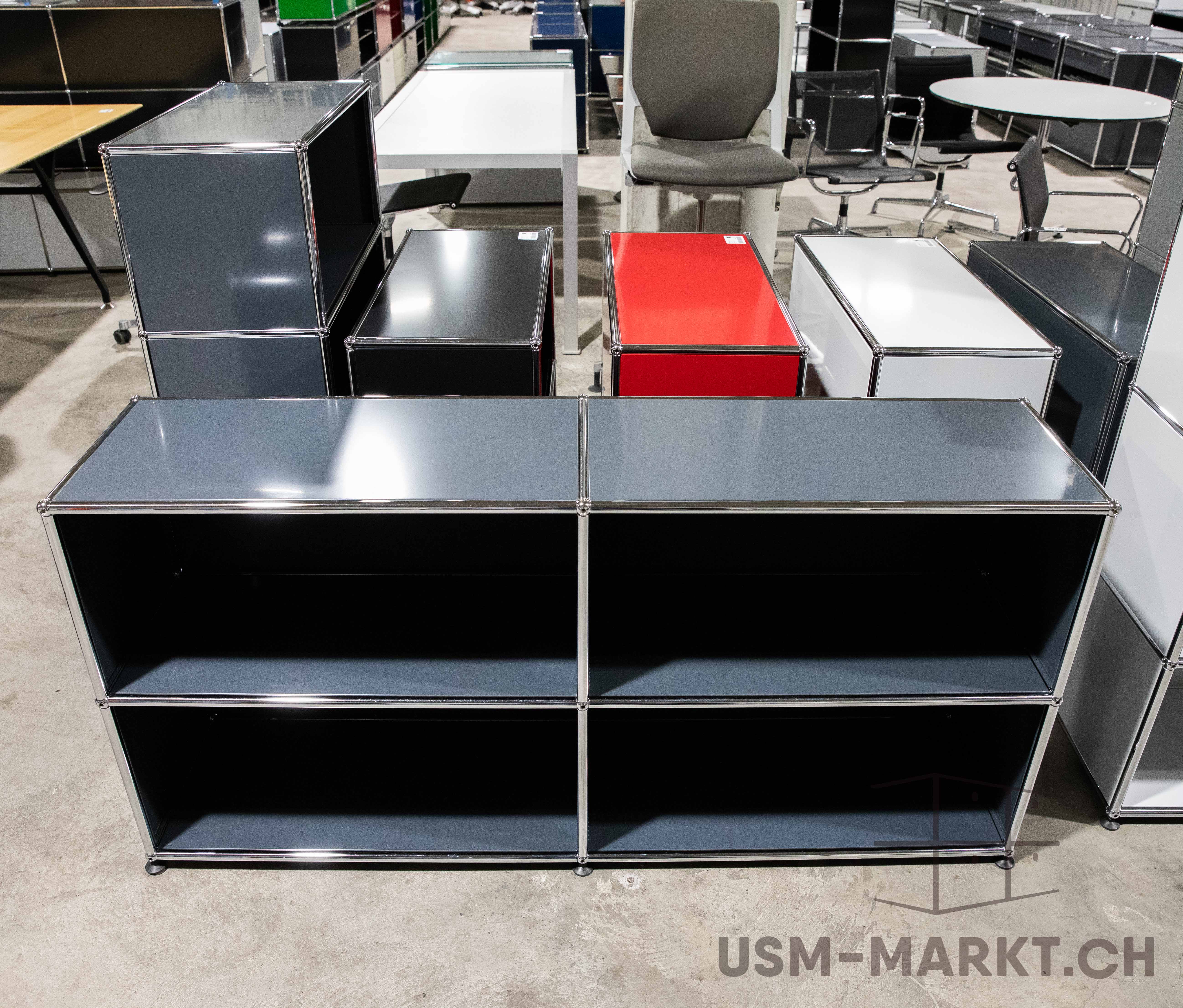 Ein graues, offenes USM Haller Sideboard mit vier Fächern steht vorn auf Betonboden. Im Hintergrund sind weitere modulare Büromöbel wie Schränke, Tische und Stühle in Grau, Schwarz, Rot und Weiss auf einer grossen Lagerfläche zu sehen.