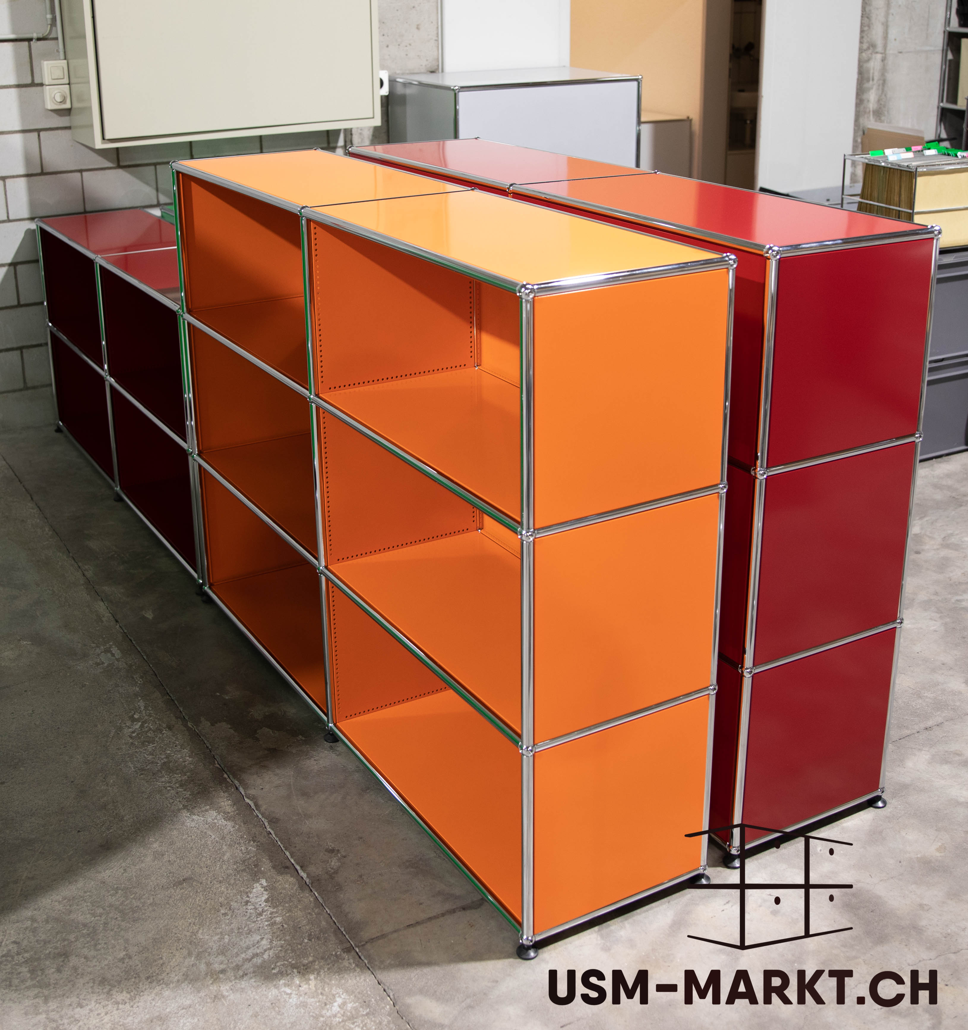 Moduli USM Haller in un ripostiglio. In primo piano due scaffali arancioni lunghi e bassi, ciascuno con tre ripiani aperti. Dietro a destra due armadi rossi più alti; a sinistra uno scaffale rosso scuro.