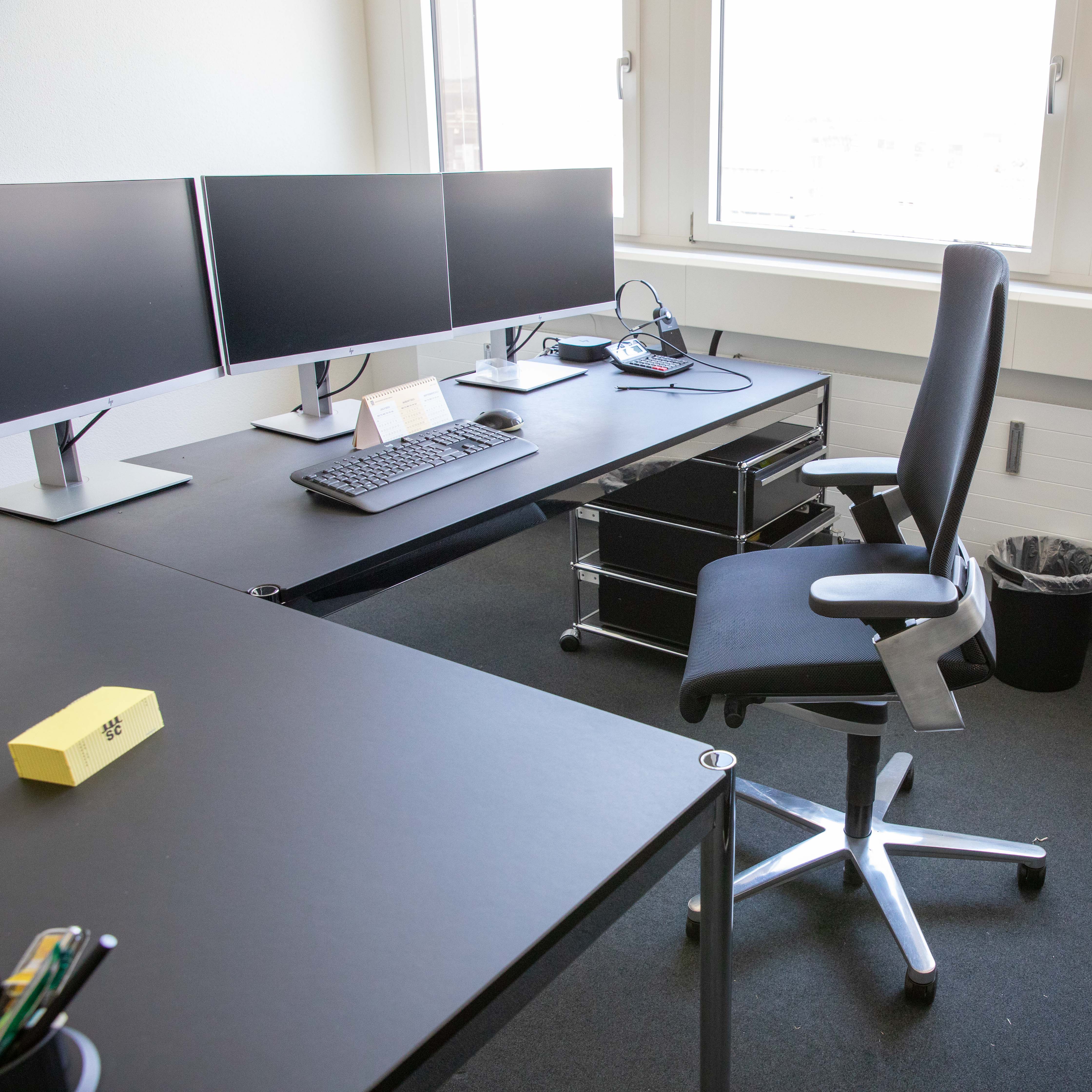 Schwarzer Wilkhahn ON Bürodrehstuhl an schwarzem L-Schreibtisch in hellem Büro. Hohe Rückenlehne, schwarze Armlehnen mit Gebrauchsspuren, silbernes Fußkreuz mit Rollen. Auf dem Tisch: 2 Monitore, Tastatur, Utensilien. Fenster hinten.