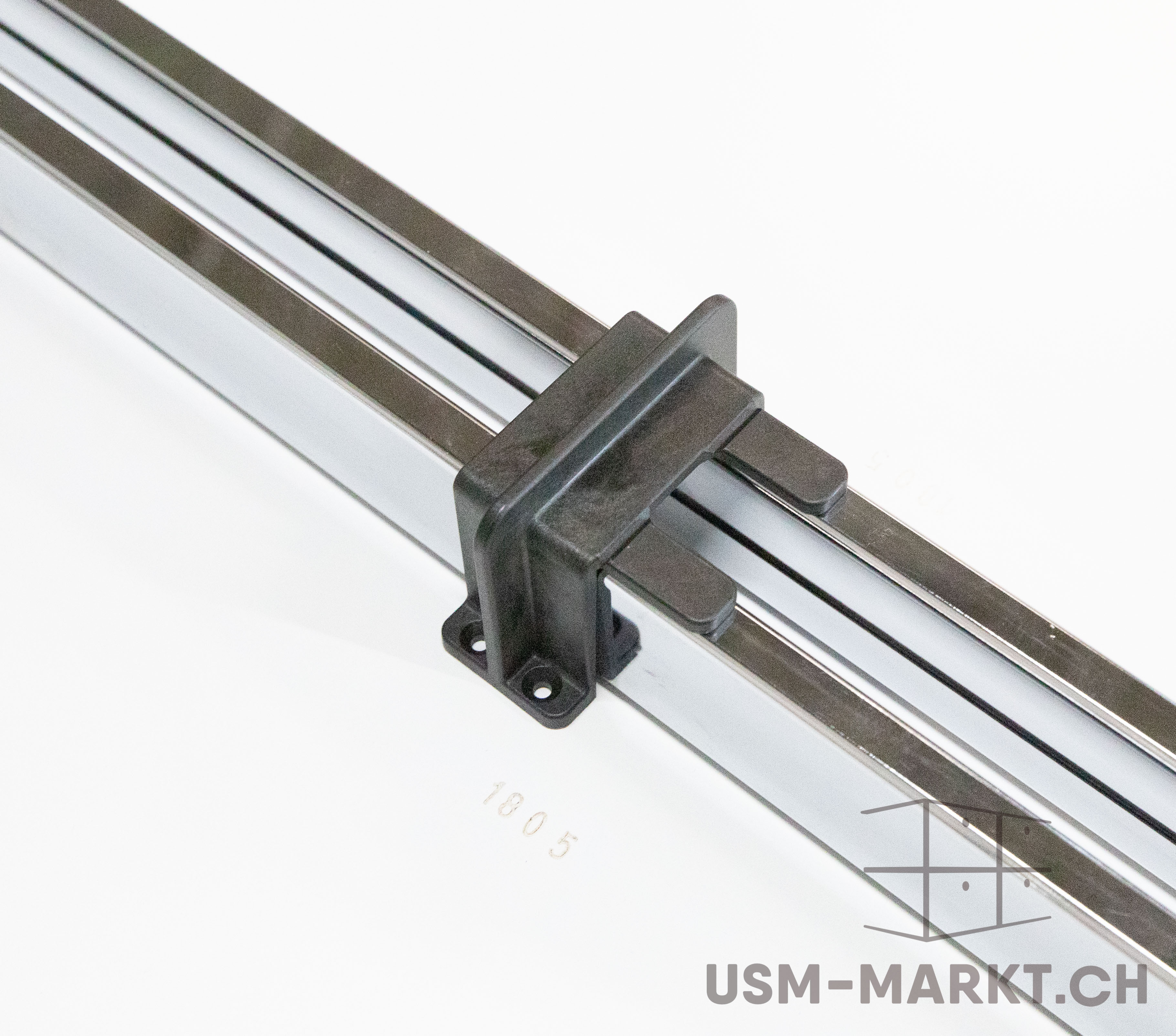 Two parallel, polished metal profiles are seen from above at an angle. A black plastic component connects them in the centre, featuring a mounting plate (2 screw holes) and two tabs above the upper profile. Light white background.
