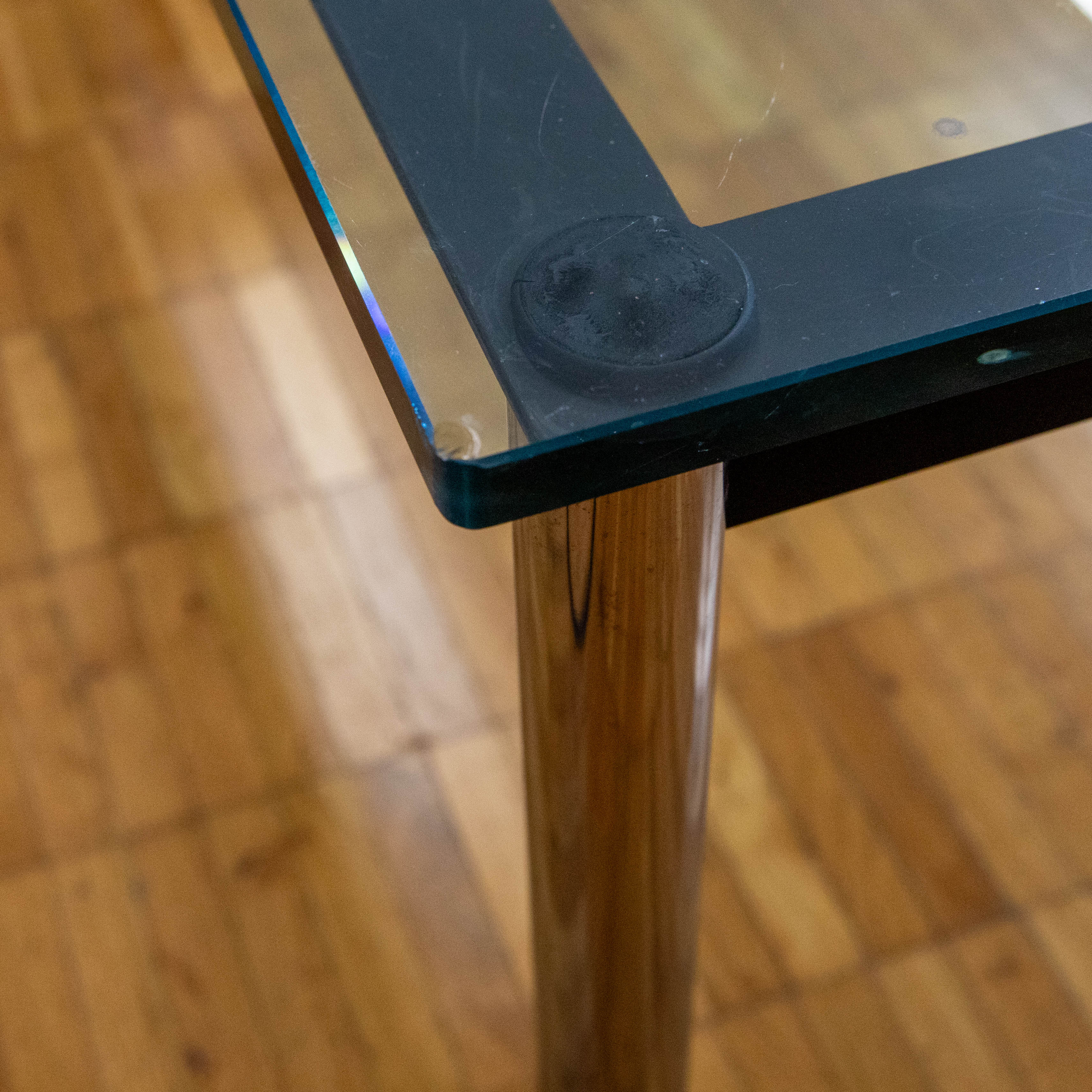 Gros plan : coin supérieur droit d'une table en verre avec un rebord métallique foncé. On aperçoit un pied de table cylindrique en métal brillant. La table repose sur un parquet clair ; la surface en verre présente des rayures et de la poussière.