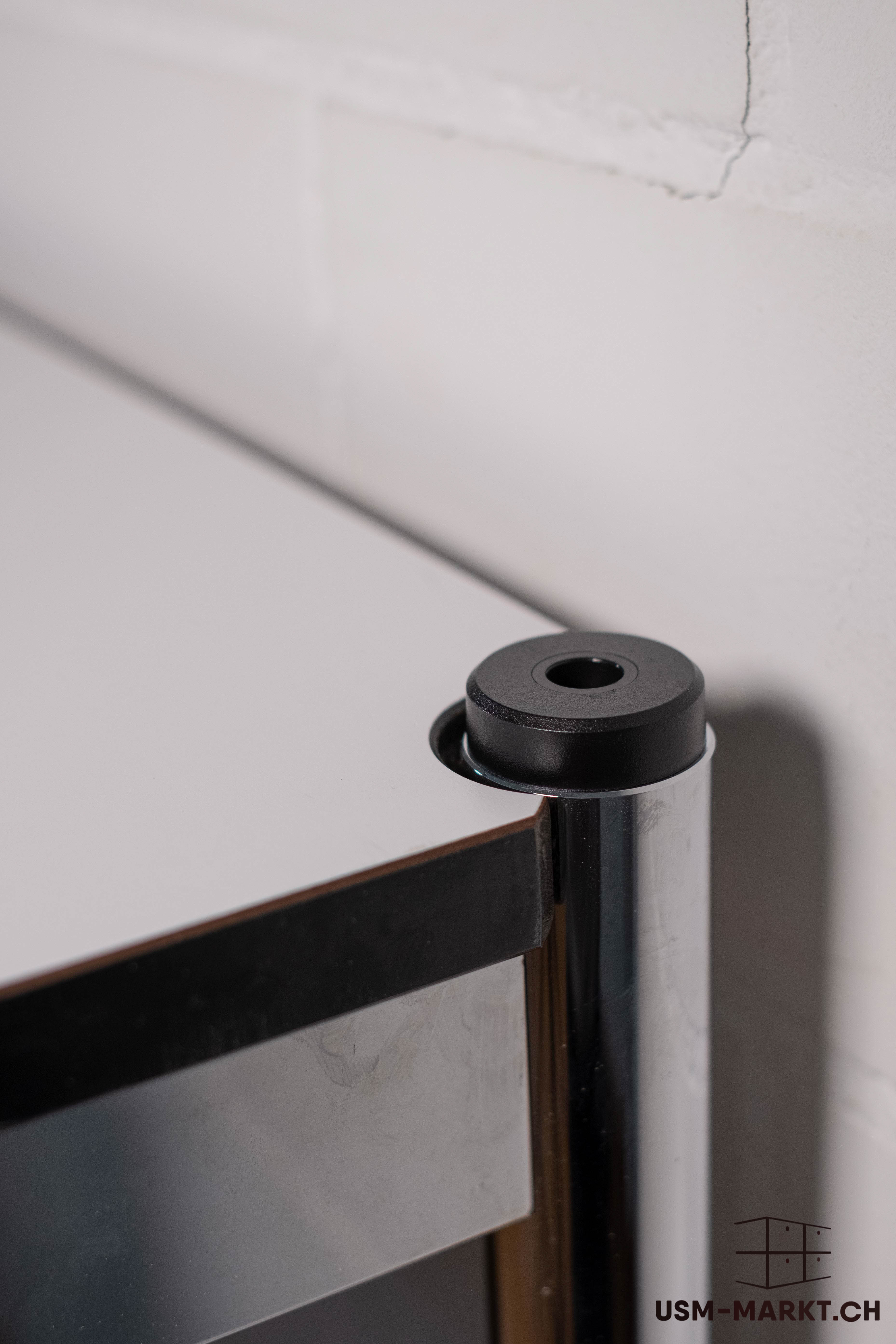 Detailed view of modern furniture: a white, smooth shelf surface next to a shiny, chrome-plated support tube with a black connecting cap. The tube shows slight wear and reflections. Out of focus: a light wall with a fine crack.