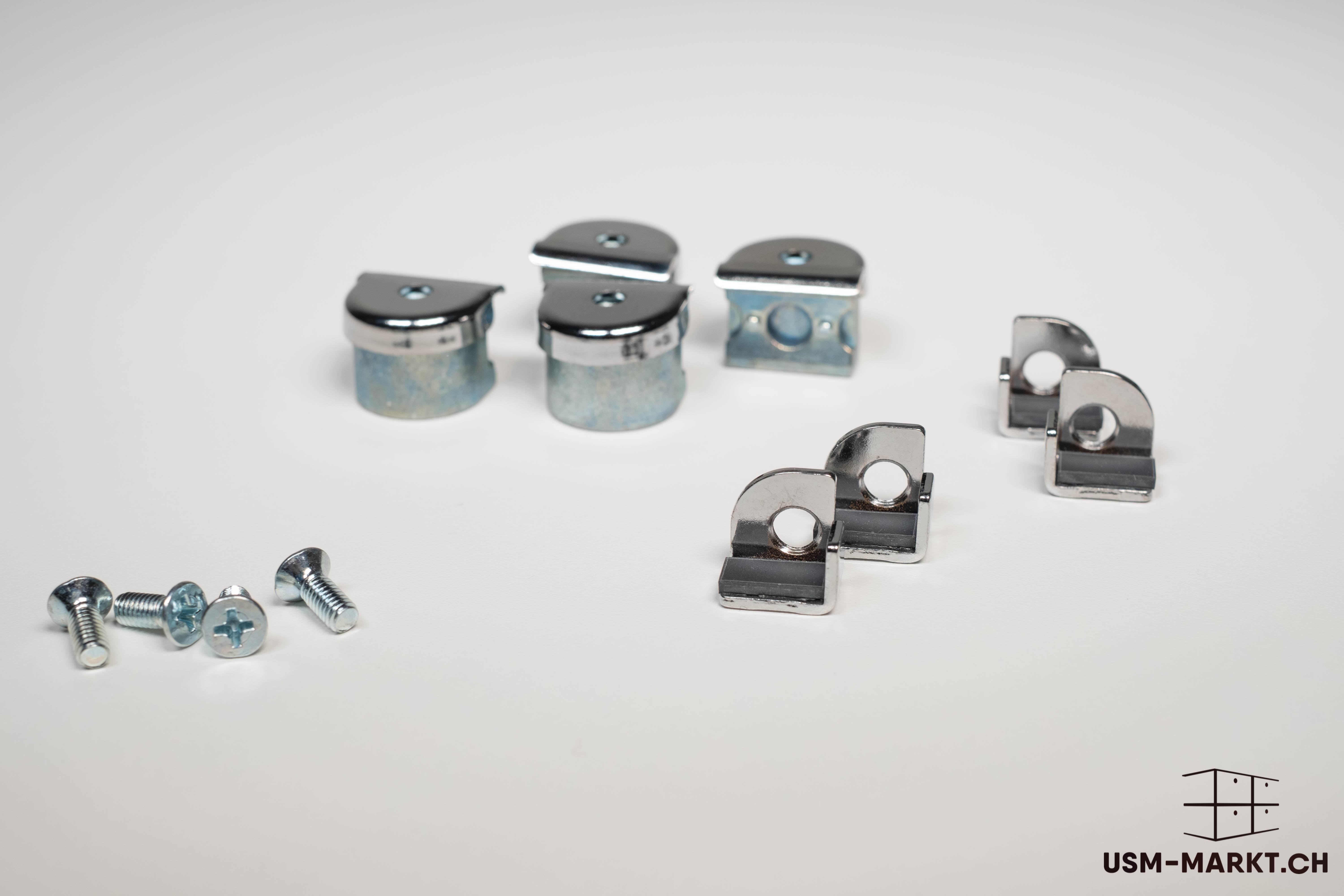 Close-up of shiny metal furniture fixings against a light grey background. At the bottom left, several small silver Phillips-head screws. In the centre and at the top, four cylindrical connectors with caps and four L-shaped corner connectors.