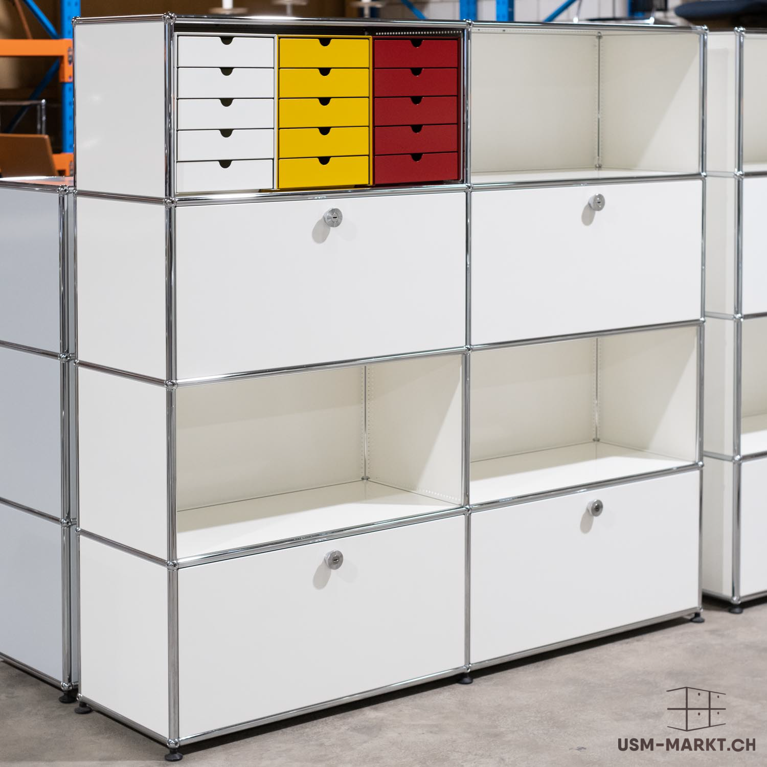 Sistema di scaffalature modulari bianco con telaio cromato, composto da diversi elementi sovrapposti. In alto a sinistra, tre file da cinque cassetti colorati ciascuna, nei colori bianco, giallo e rosso. Sotto, armadi chiusi e ripiani aperti.