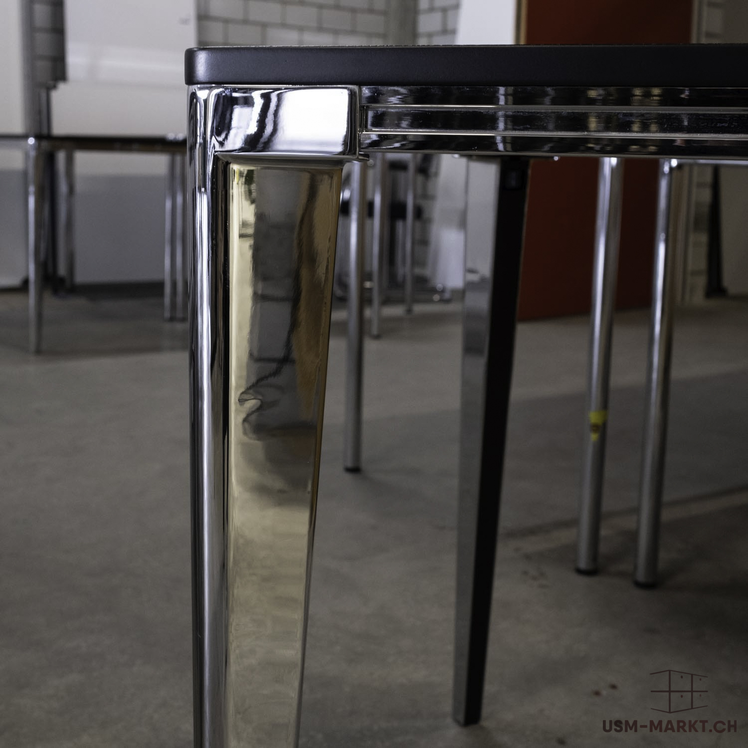 Gros plan sur un coin de table avec un plateau noir et un pied en chrome brillant. Le pied poli reflète les autres meubles et un mur rouge en arrière-plan. Le sol en béton gris met en valeur le design moderne et fonctionnel.