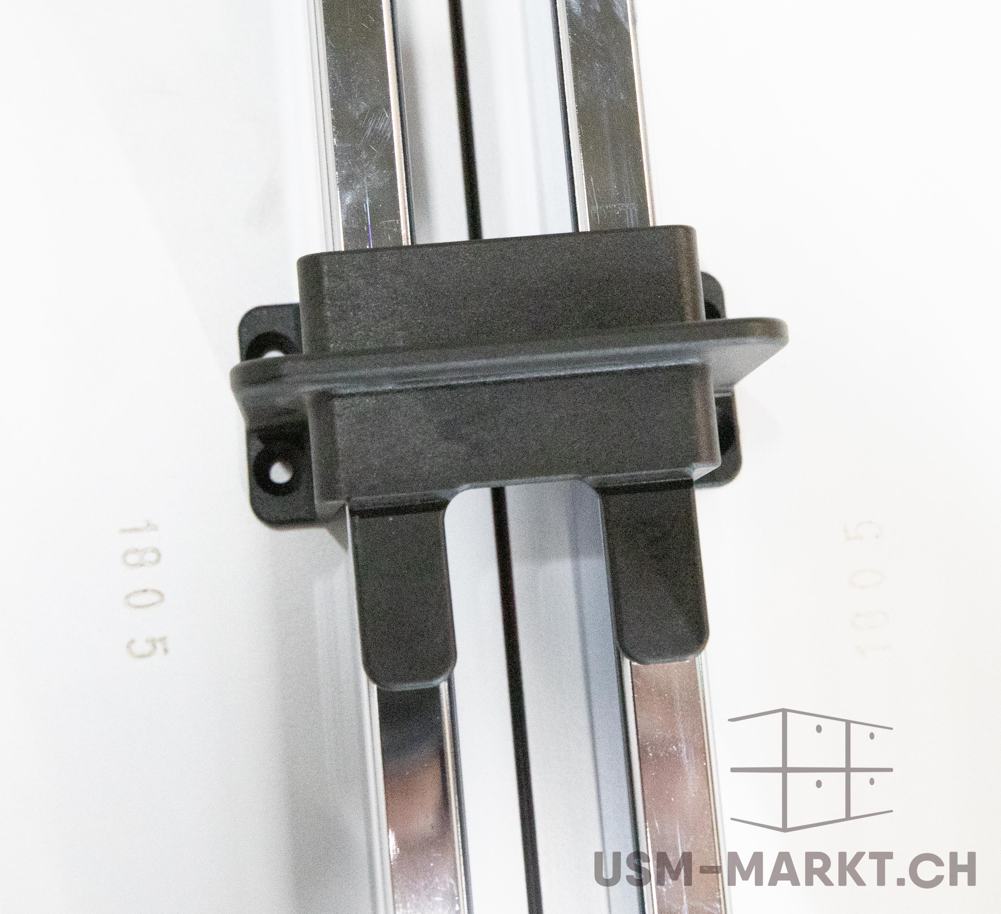 A black plastic connector secures two vertical, shiny metal rods against a white background. The sturdy connector encases the parallel, slightly scratched rods. The numbers ‘1805’ and ‘1005’ are visible in the background.
