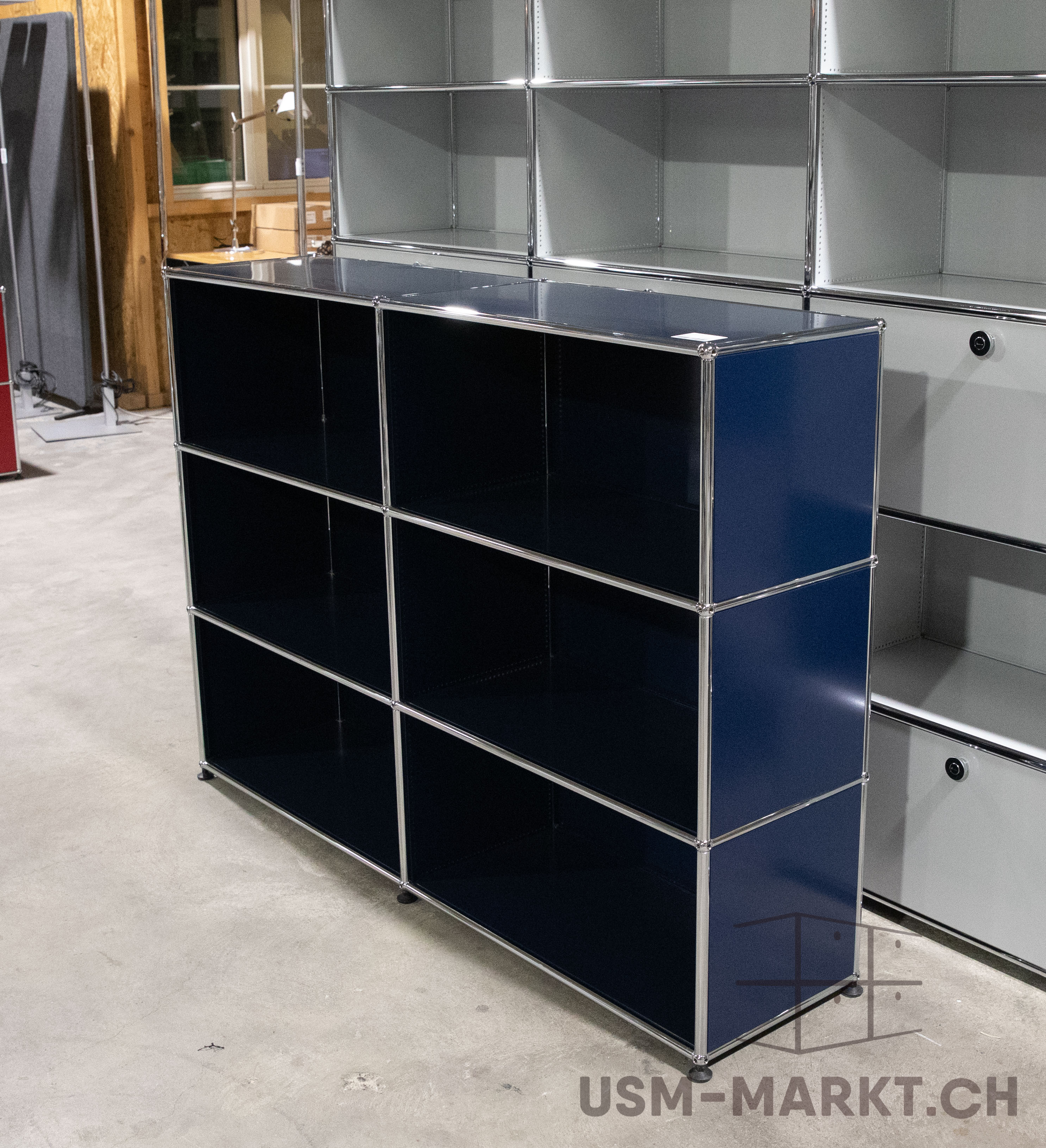 Scaffale USM Haller blu scuro con sei ripiani aperti e telaio cromato su pavimento in cemento grigio chiaro. Dietro, altri scaffali modulari grigio chiaro e in parte rossi, in ambiente magazzino/espositivo.