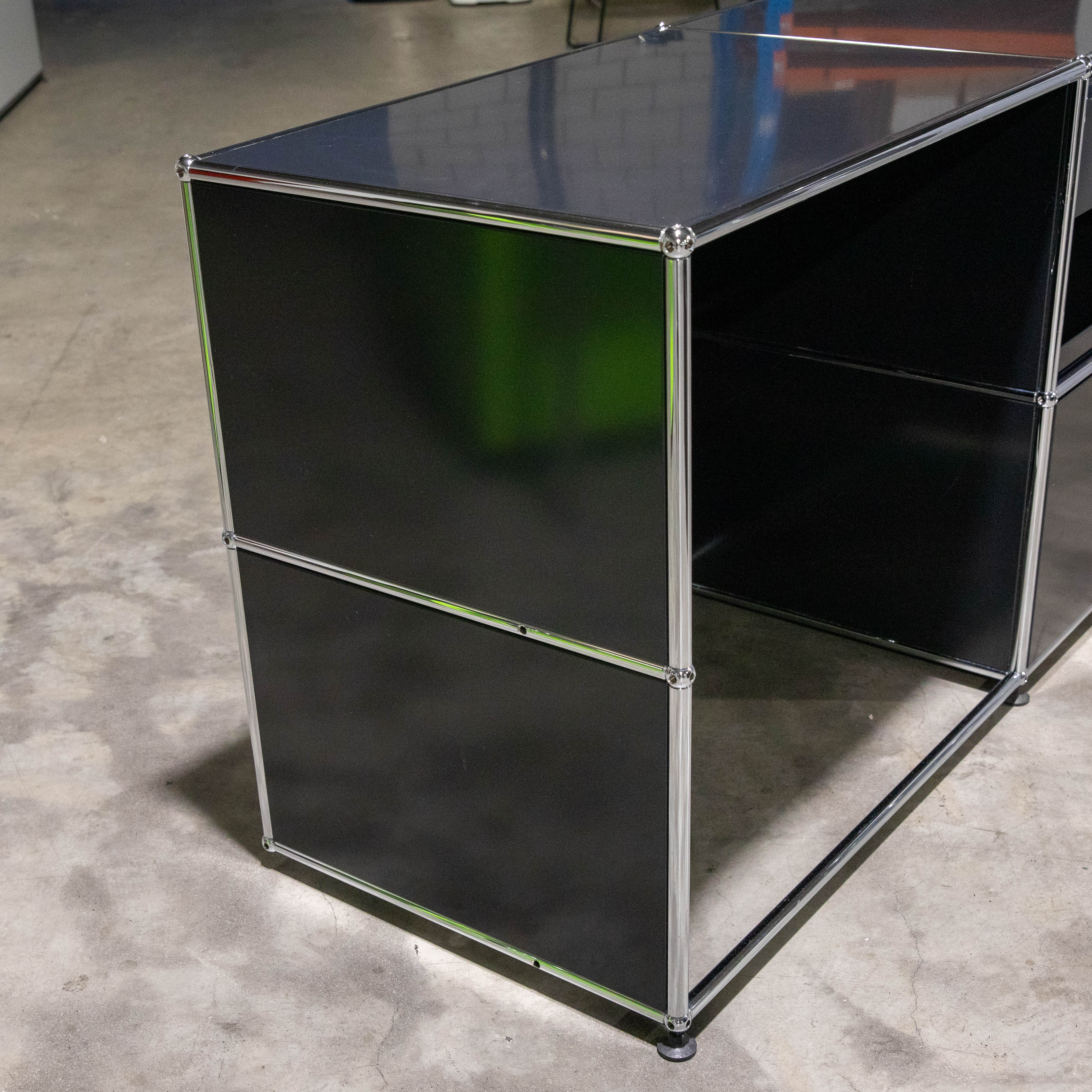 Schwarzes modulares Möbel mit glänzenden Chromrahmen und Verbindern auf hellgrauem Betonboden. Links zweistöckig mit dunklen Paneelen, rechts offen als Schreibtisch- oder Regalstruktur. Grüne Reflexionen sichtbar.