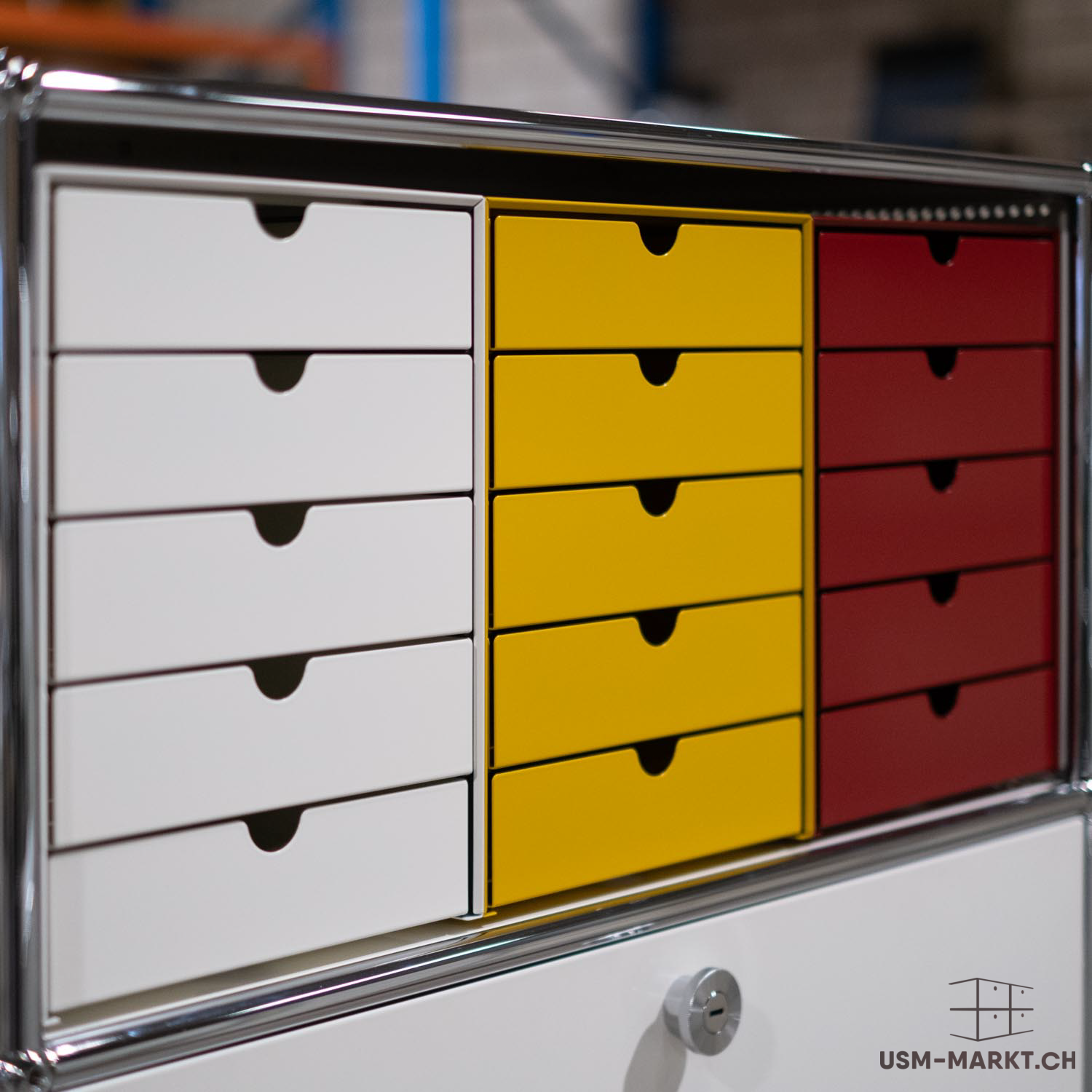 Credenza USM Haller con tre colonne di cassetti: la prima bianca, la seconda gialla, la terza rossa. Ogni colonna ha sei cassetti con maniglie incassate semicircolari. Acciaio cromato lucido. Nella parte inferiore, ripiano bianco con serratura a cilindro.