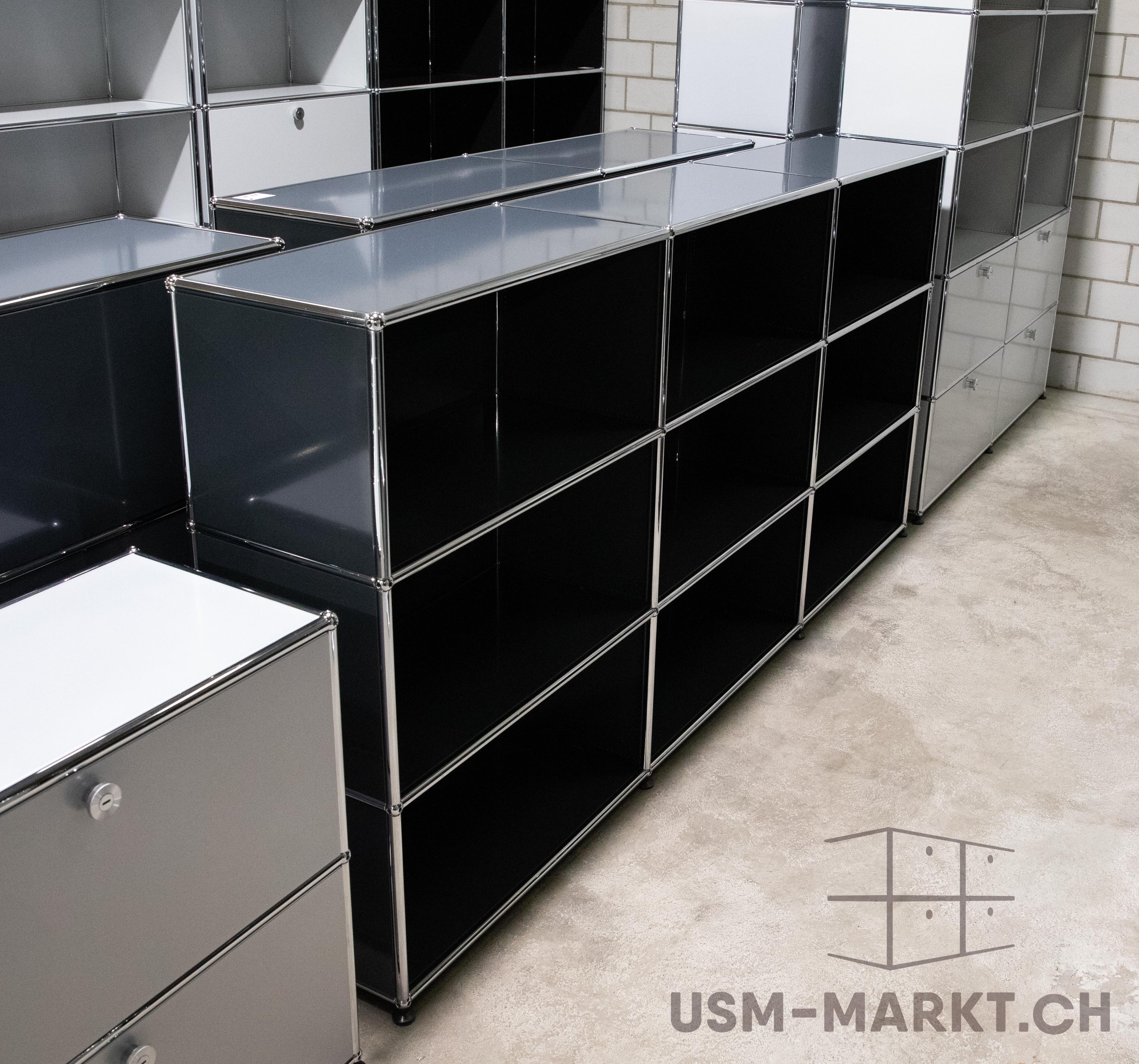 Reihen von USM Haller Möbeln in einem Lagerraum: glänzend schwarze, weiße und graue Module mit Chromrahmen. Links weiße und graue Schubladen, rechts hohe Regale an einer weißen Ziegelwand. Heller Betonboden.