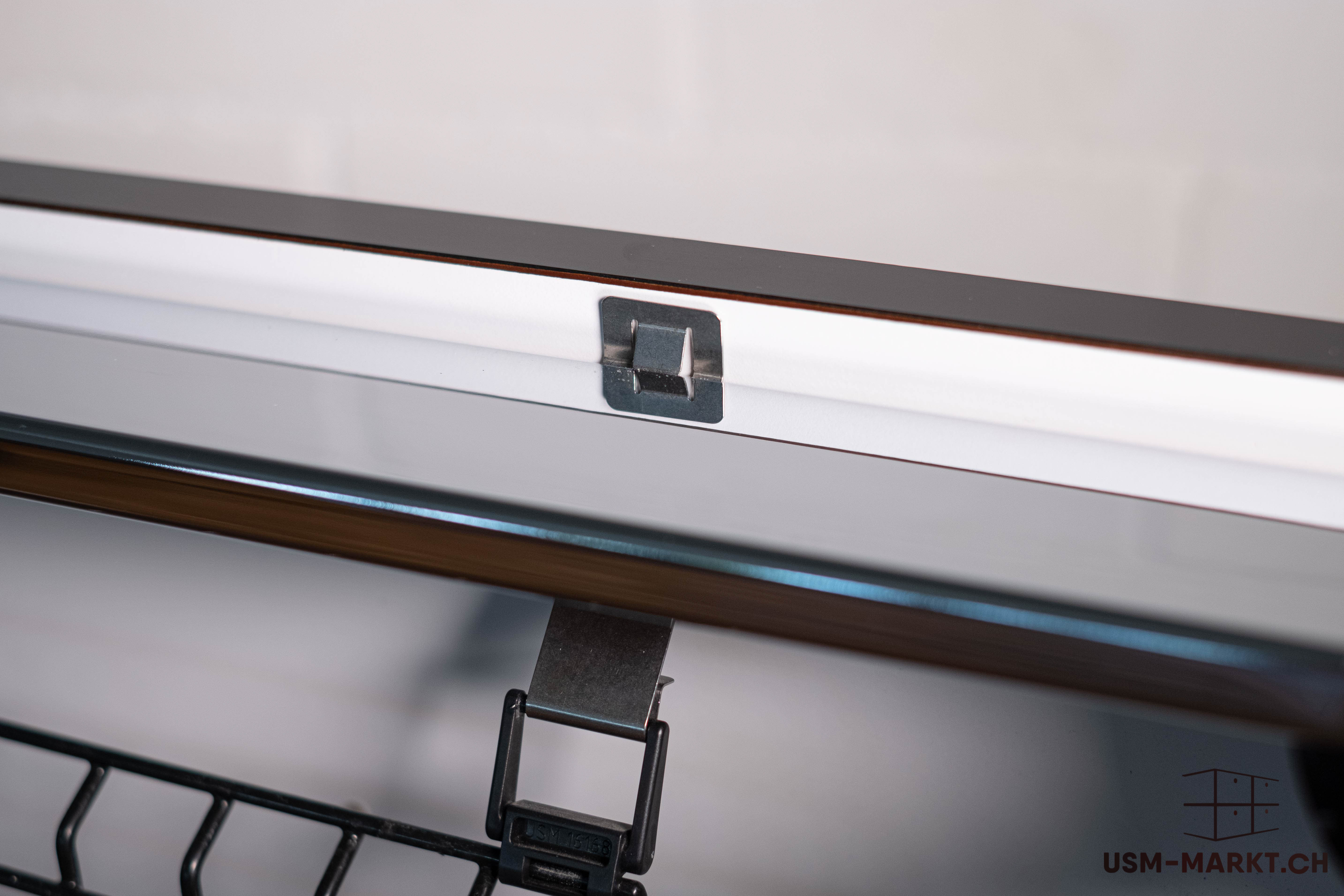 Nahaufnahme der oberen Innenkante eines Möbelstücks, vermutlich USM Haller. Ein glänzendes Metallprofil verläuft horizontal, darüber ein dünner dunkler Streifen. Am oberen Rand sitzt ein kleiner Metallclip. Darunter eine schwarze Gitterstruktur.