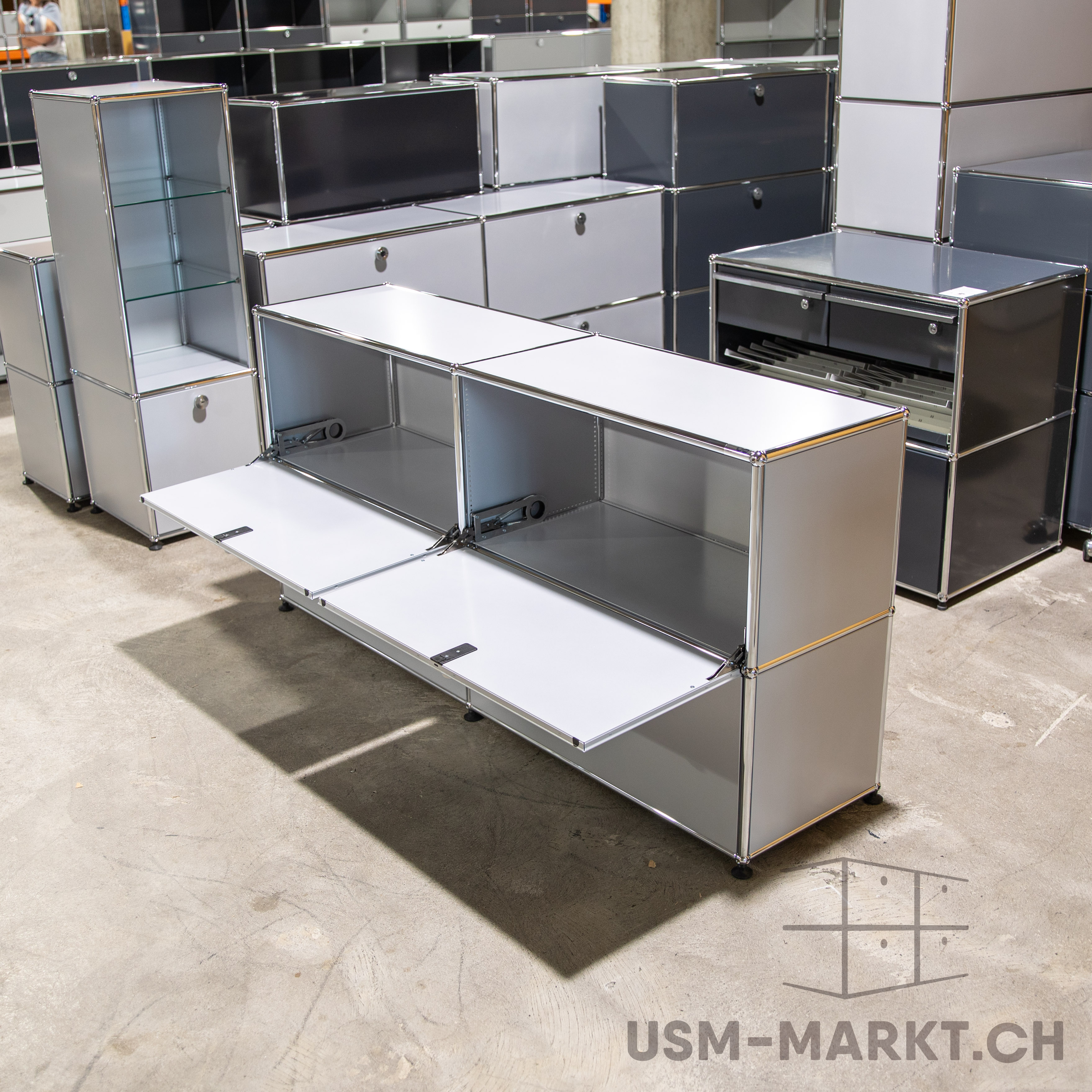 Lagerraum mit modularen USM Haller Möbeln. Vorn ein langes weißes Sideboard mit zwei offenen Klapptüren, daneben ein hohes schmales silbernes Regal mit Glasböden. Hinten weitere USM-Schränke und -Regale in Grau/Weiß.