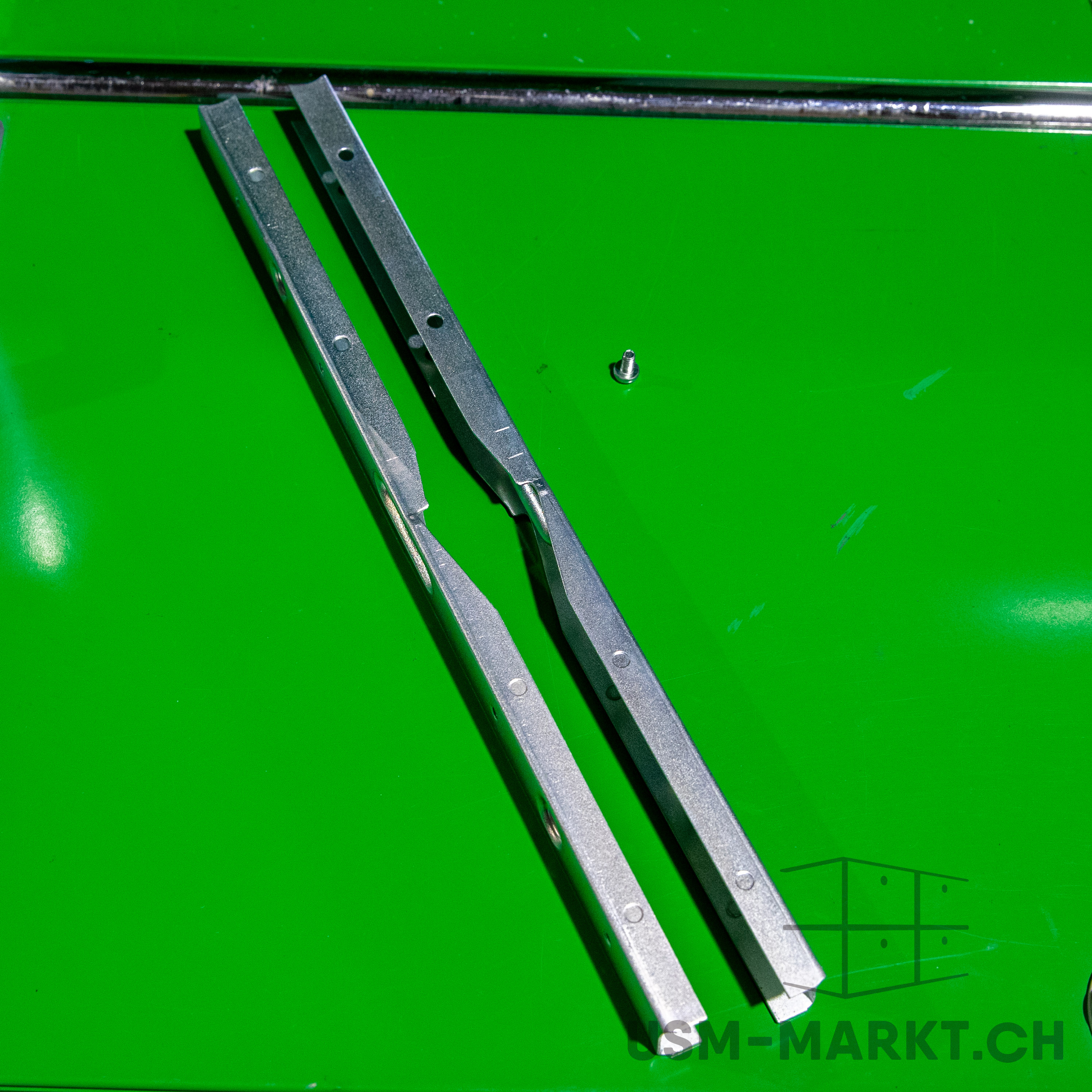 Two silver-coloured, U-shaped metal rails lie parallel to one another on a shiny, bright green surface. Each rail has several small round holes. To the right of the rails lies a single small flat-headed screw.