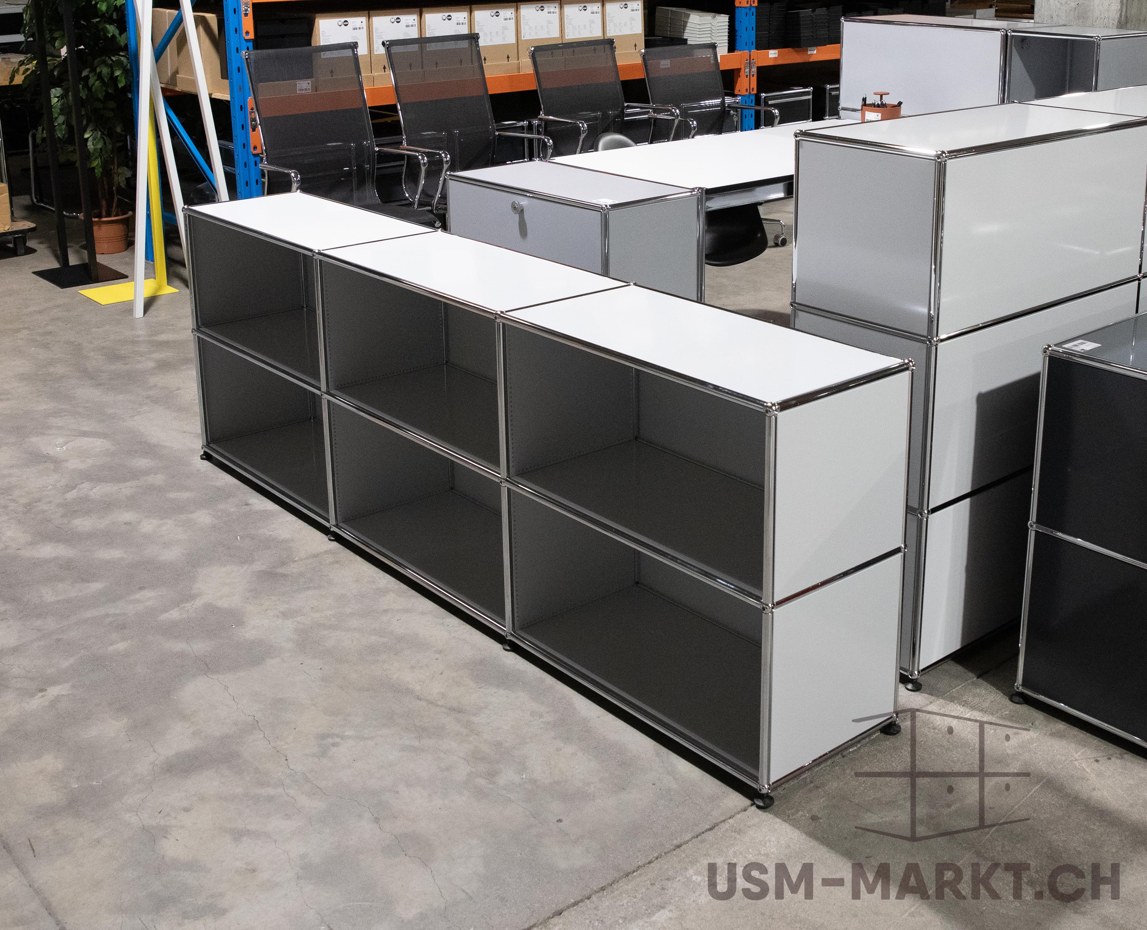 Lagerraum mit modernen, modularen Möbeln: Vorn langes weißes USM Haller Regal mit 8 offenen Fächern und Chromrahmen. Dahinter weitere USM Haller Sideboards, Schreibtisch mit Bürostühlen, Industrieregale mit Kartons und Pflanze. Betonboden.