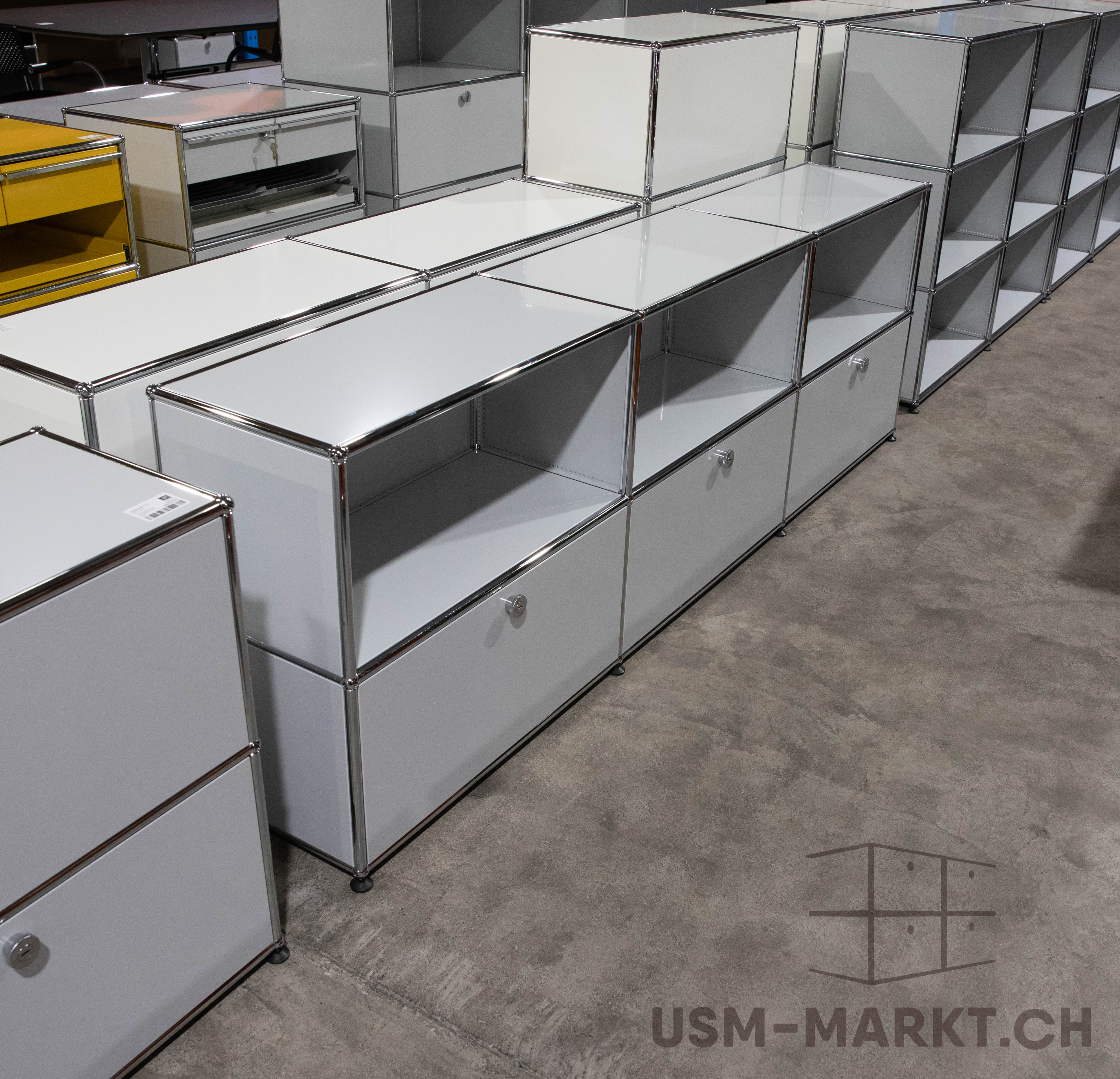 Weiße und hellgraue USM Haller Möbel füllen einen Großteil des Raumes, in verschiedenen Konfigurationen als Sideboards, Regale und Schränke. Einige mit offenen Fächern, andere mit Schubladen, Griffen oder Schlössern. Links ein gelber Schrank.