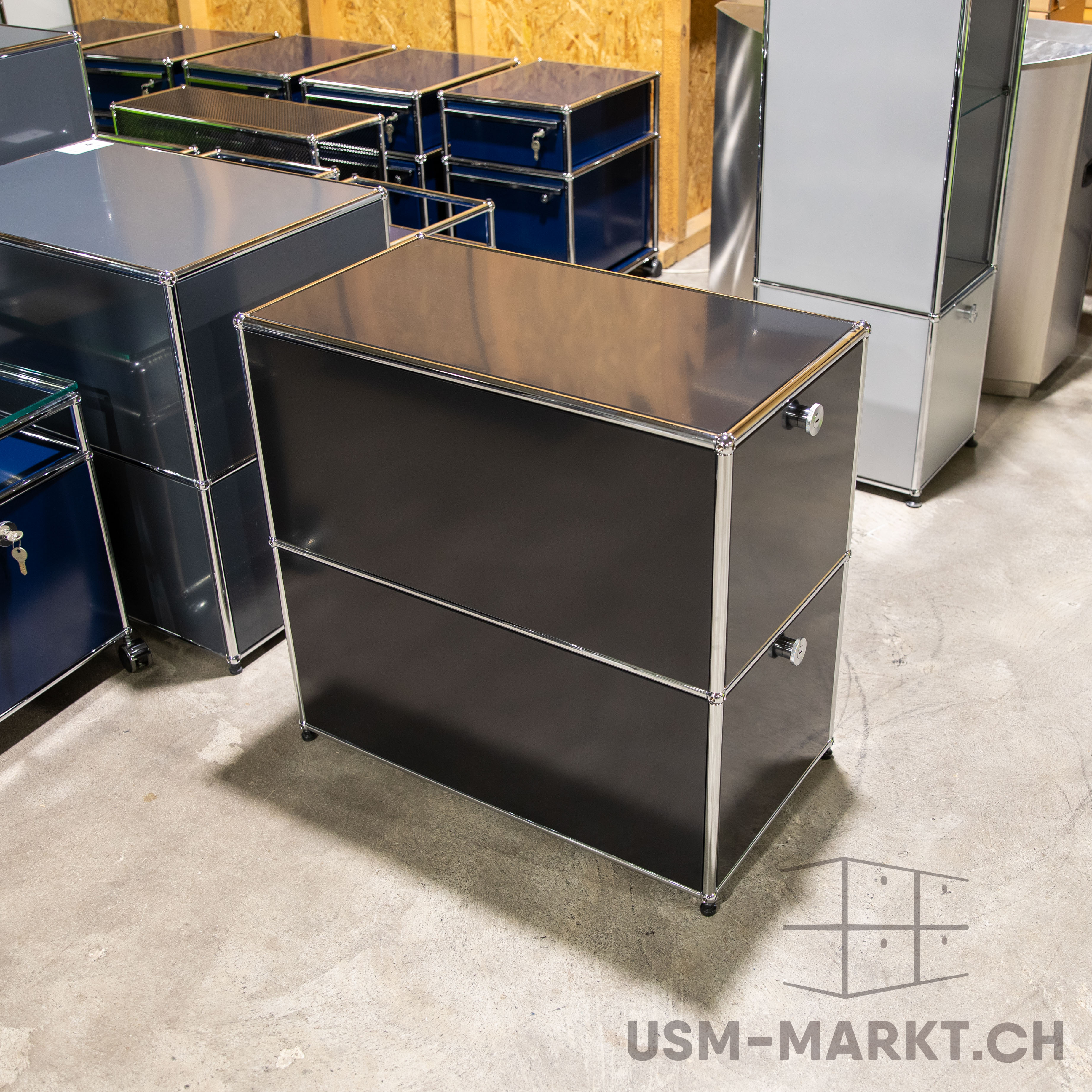 USM Haller Möbel in einem Lagerraum. Vorn ein anthrazitfarbenes Sideboard mit zwei Schubladen. Dahinter weitere dunkle und blaue USM-Module, teils mit Schlüsseln. Rechts ein höheres, hellgraues Regal. Betonboden.