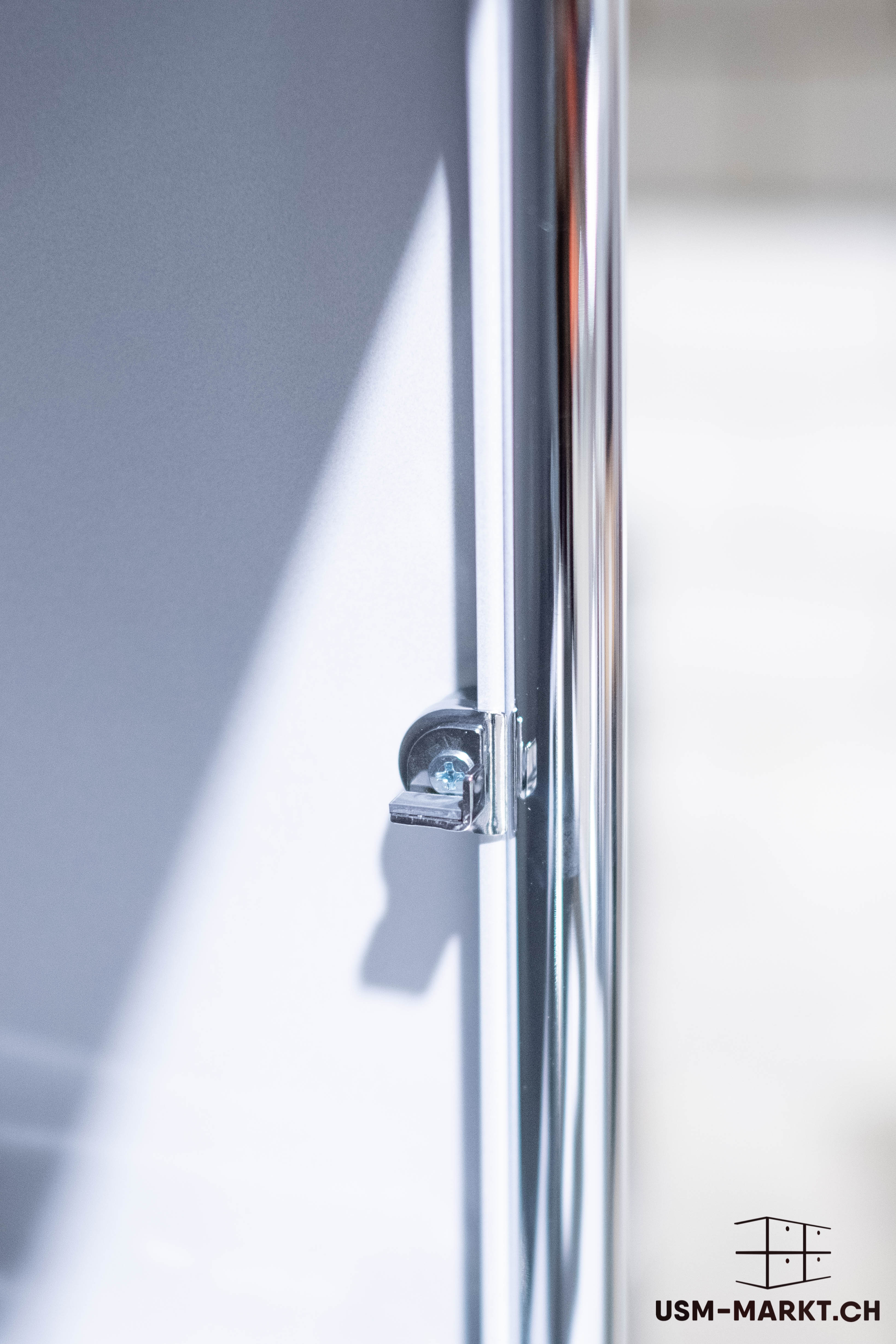 Close-up of a shiny chrome tube on the right-hand edge of the image. On the left, a light grey/white furniture panel, secured to the tube with a small L-shaped metal fitting and a screw. Reflections and highlights on the metal.
