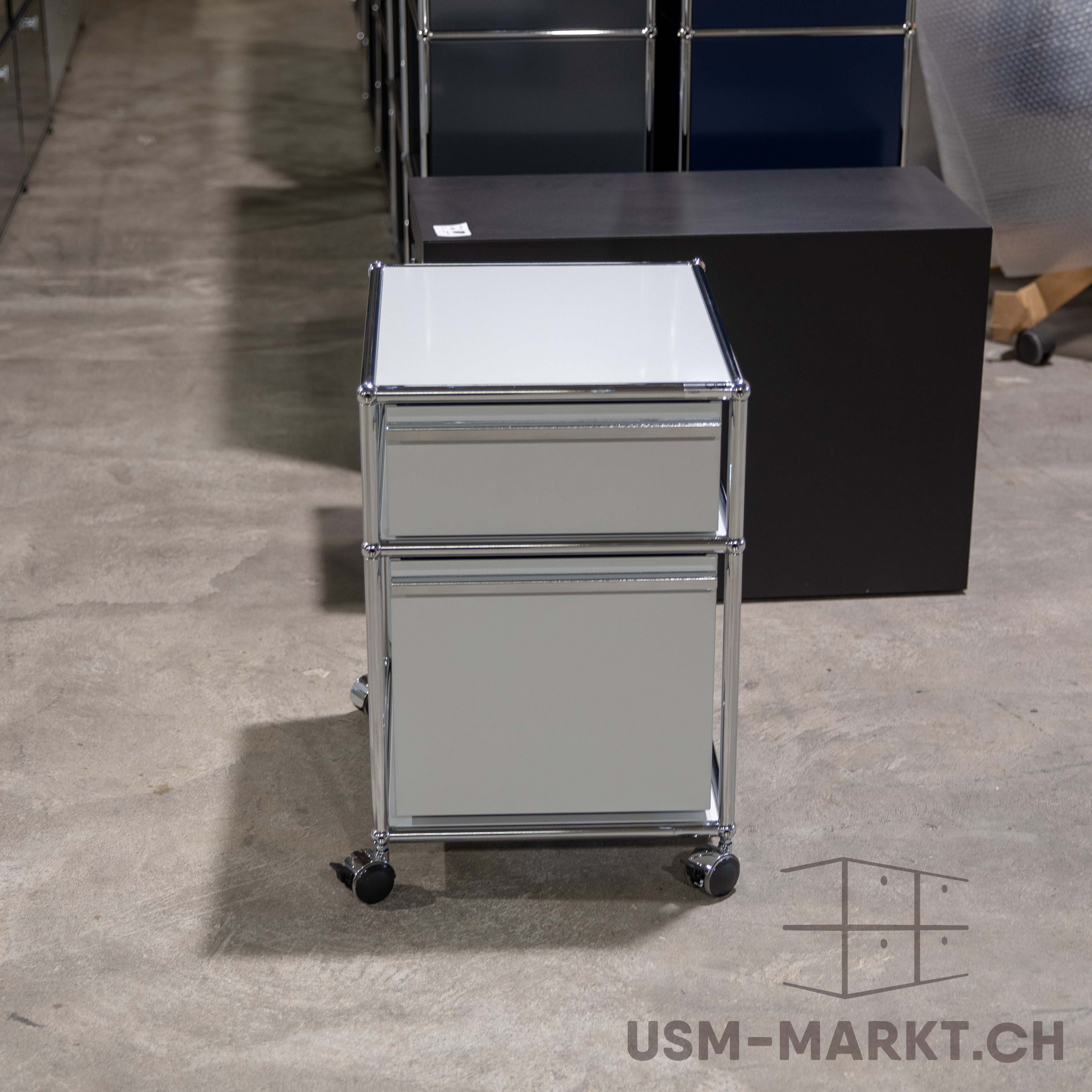 Hellgrauer, minimalistischer Rollcontainer mit zwei geräumigen Schubladen und Chromgestell. Steht auf vier schwarzen, leichtgängigen Rollen auf Betonboden. Im Hintergrund unscharfe dunkle Möbel/Regale, wie Lager oder Ausstellung.
