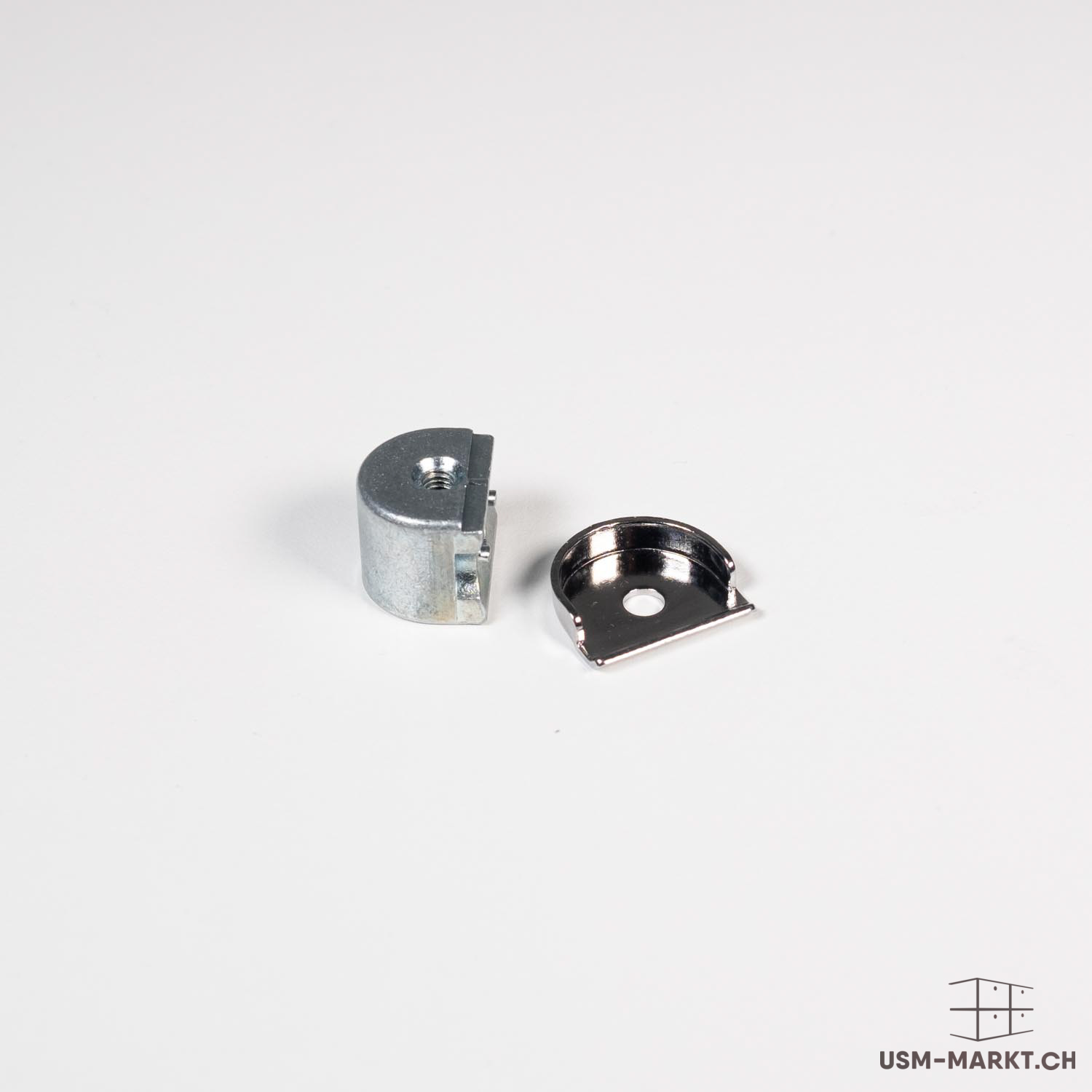 Two metal furniture connectors on a white surface. On the left, a cylindrical, silver cast component with an internal thread, similar to an eccentric connector. On the right, a smaller, shiny U-shaped component with a round opening.