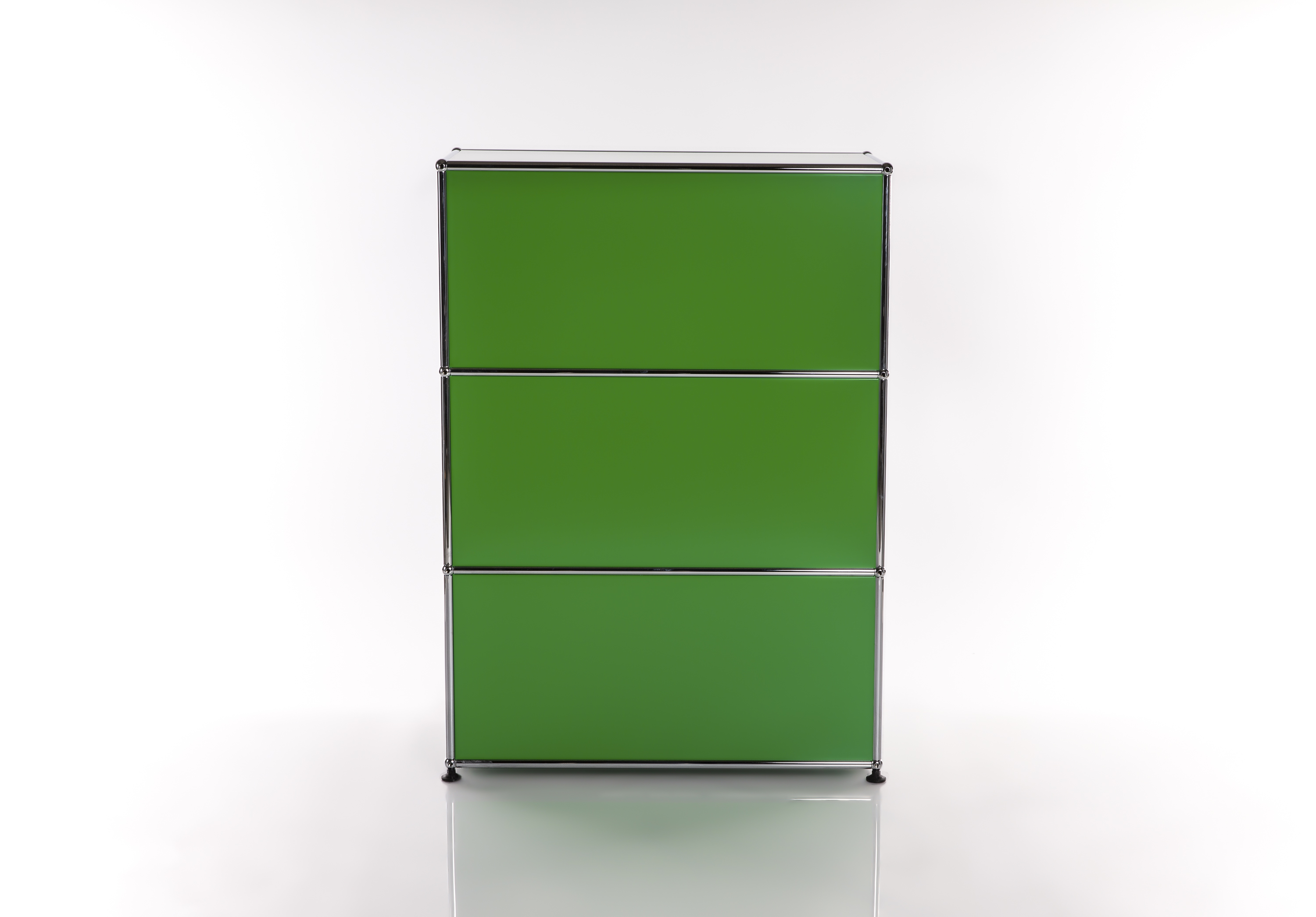 A sleek, modular shelving system in bright green, consisting of three stacked compartments. The fronts are matt green, framed by polished chrome. It stands on a reflective white surface and is shown from the front.