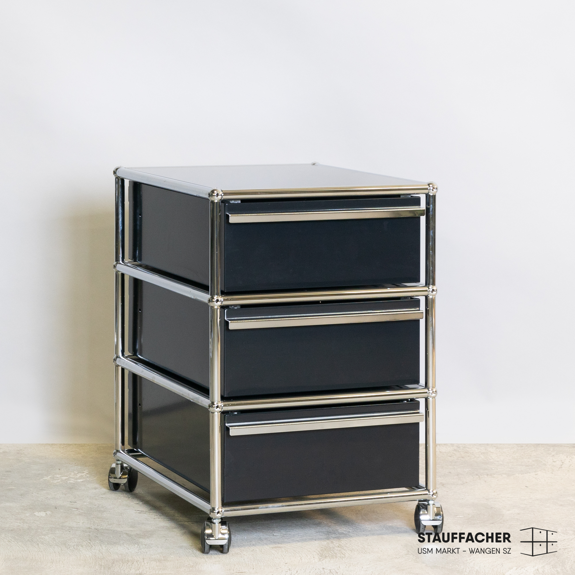 Moderner, eleganter Rollcontainer mit drei schwarzen Schubladen und glänzendem Chromrahmen. Matt-schwarze Fronten mit schlanken silbernen Griffen. Auf vier feststellbaren Rollen, auf hellem Beton vor weisser Wand.