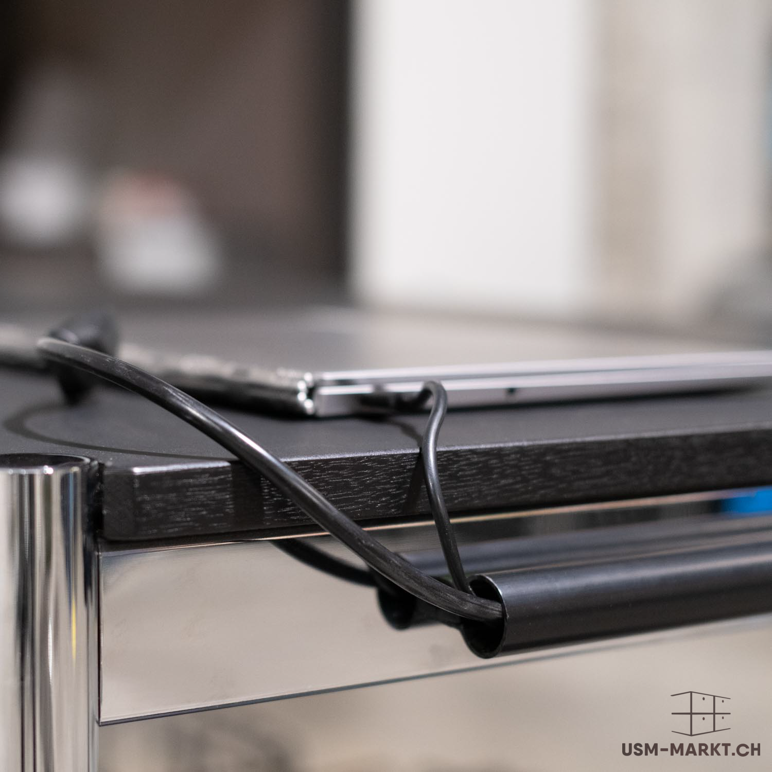 Nahaufnahme einer schwarzen Tischplatte aus Holz, auf der ein silberner Laptop und ein schwarzes Kabel liegen. Das Kabel ist sauber durch eine schwarze Kabelkanal-Röhre unterhalb der Platte geführt. Links sind glänzende Chromstangen sichtbar.