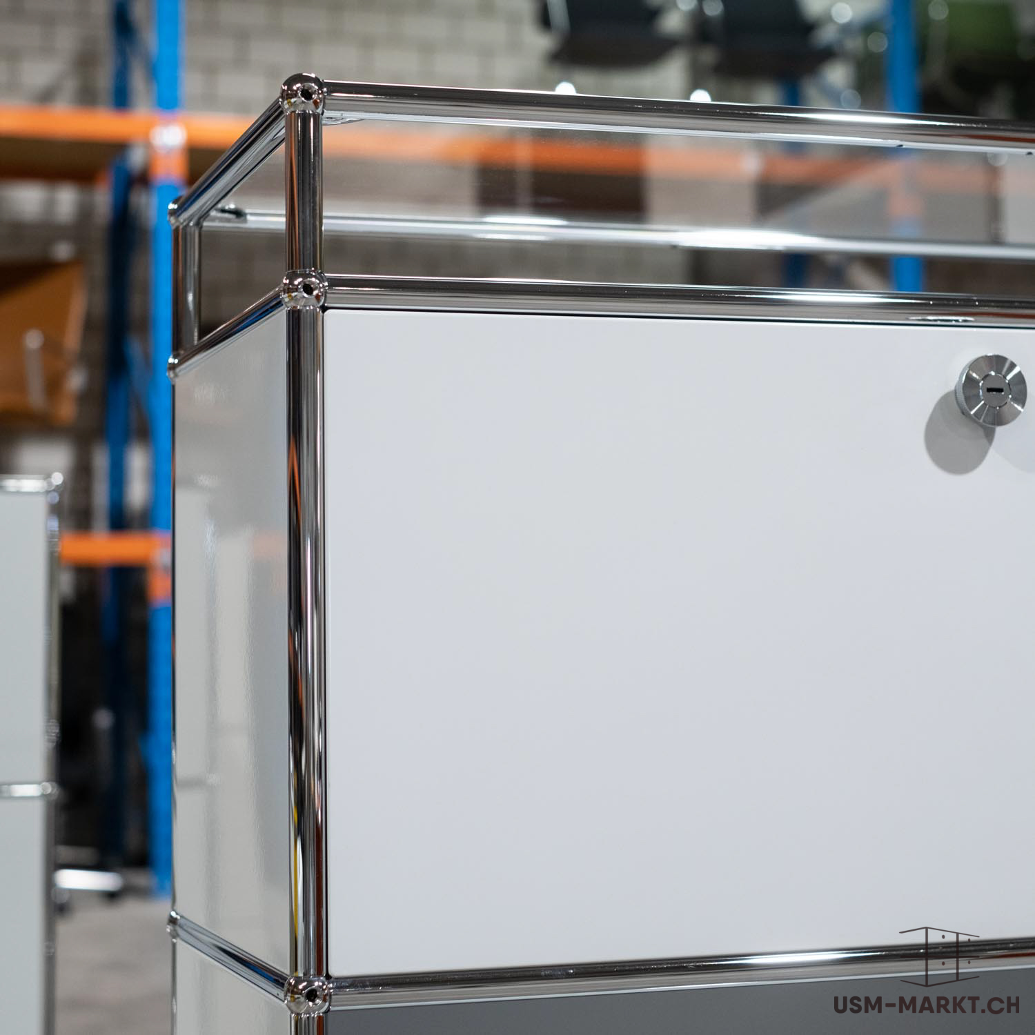 Mobile modulare USM Haller bianco con tubi cromati lucidi e giunti sferici. Piano in vetro trasparente nella parte superiore e pannelli bianchi. Chiusura argentata sul frontale bianco. Sfondo sfocato: scaffali di magazzino.