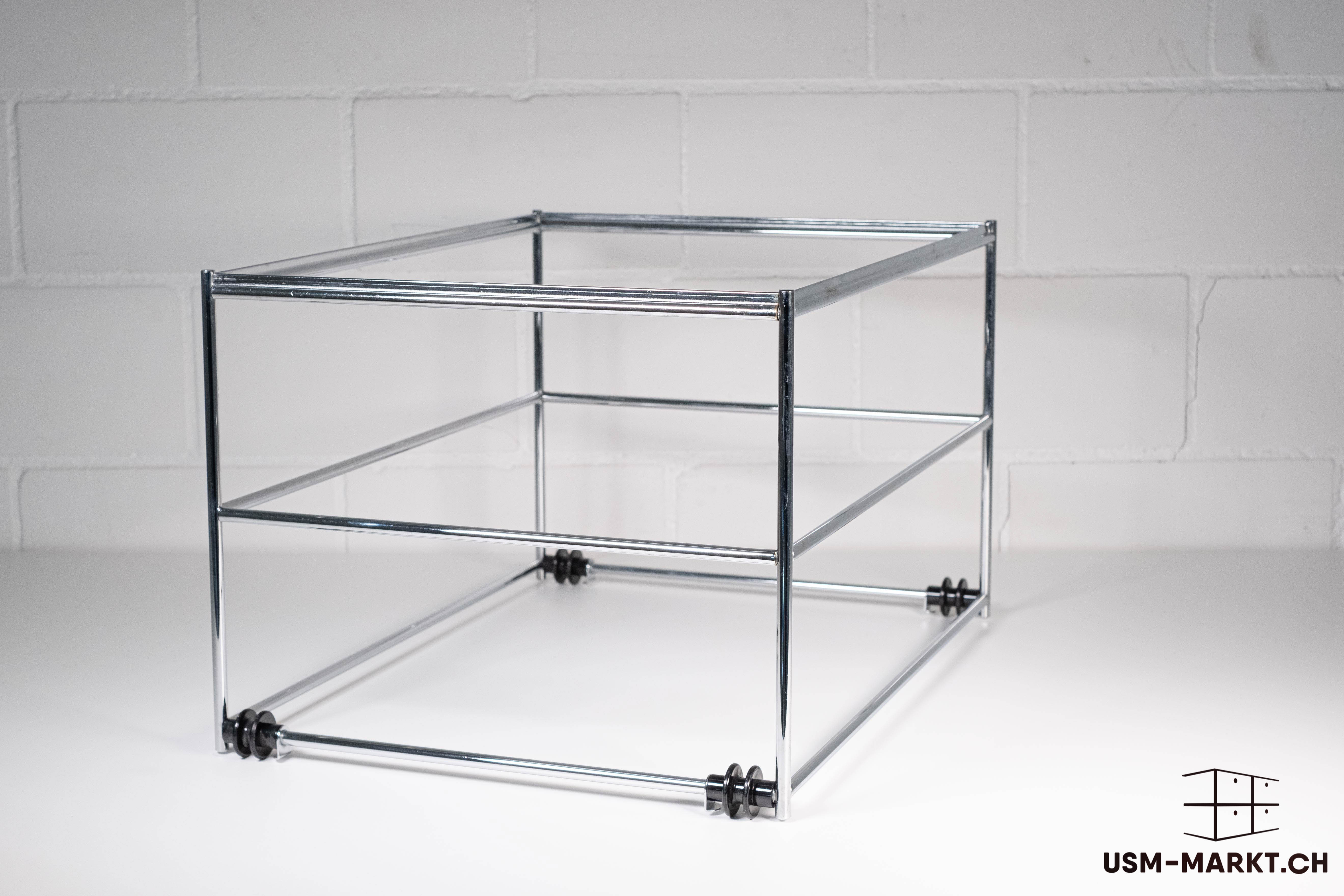 Struttura base USM Haller lucida e vuota, realizzata con tubi metallici cromati e connettori, a tre livelli e aperta, a forma di cubo. Elementi in plastica nera agli angoli inferiori. Su una superficie chiara davanti a una parete di mattoni bianchi.