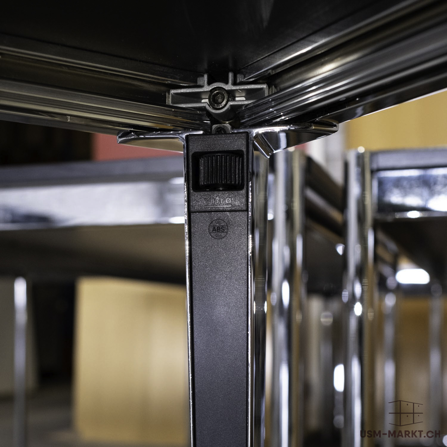 Gros plan sur la face inférieure d'un plateau de table noir sur un piètement en chrome brillant. Un élément de fixation rectangulaire noir, avec les inscriptions en relief « Vitra » et « ABS », stabilise la structure. Arrière-plan flou.