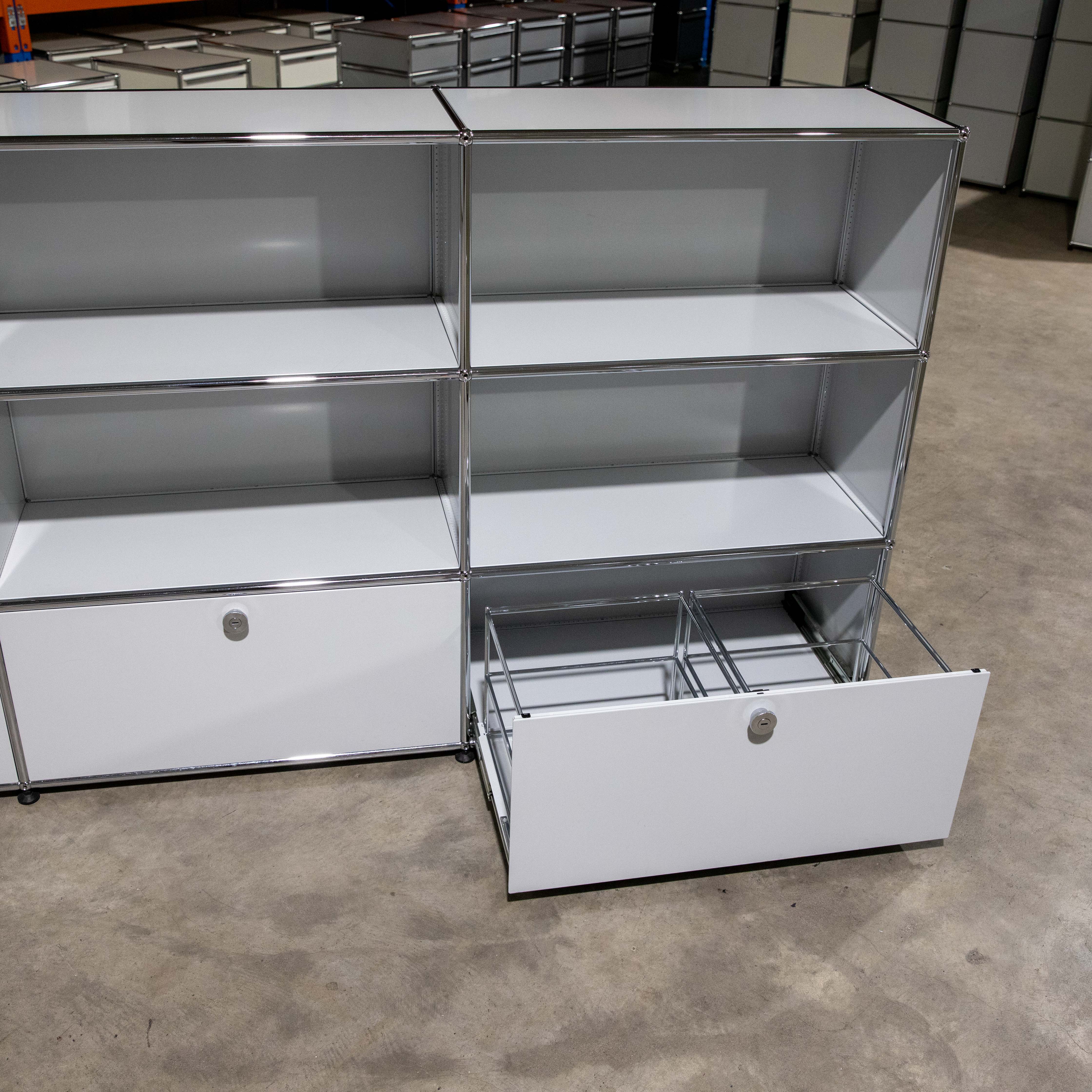 Gebrauchtes USM Haller Regal in Lichtgrau mit neun Modulen (3x3). Oben sechs offene Fächer. Unten zwei geschlossene und ein teils offenes Auszugfach mit Metallrahmen für Hängeregistratur. Steht auf grauem Betonboden.