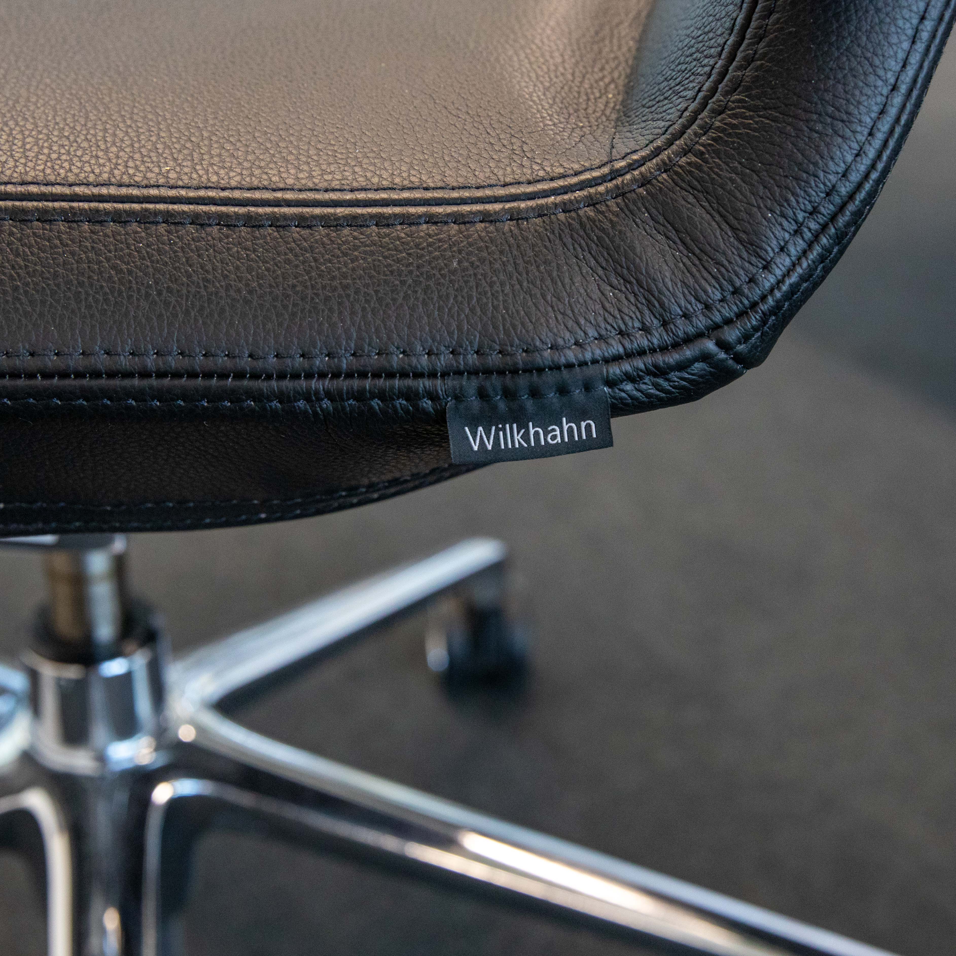 Detailansicht der schwarzen Lederpolsterung eines Bürodrehstuhls mit sichtbaren Nähten. Ein kleines schwarzes Etikett mit weißem "Wilkhahn"-Logo ist an der Unterseite des Sitzes angebracht. Im Hintergrund der Metallfuß.