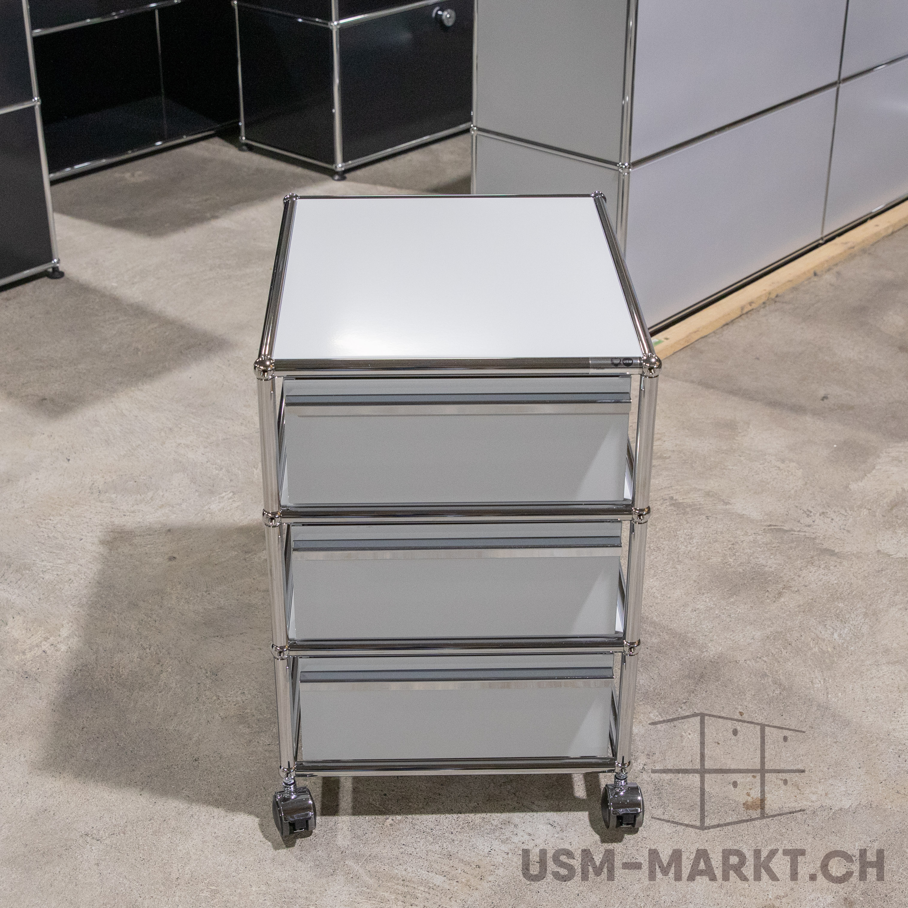 Dreistöckiger, mobiler Rollcontainer im modernen Design mit weißer Oberplatte und drei weißen Schubladen. Konstruktion aus glänzenden, verchromten Metallrohren und -verbindern. Steht auf hellgrauem Betonboden.