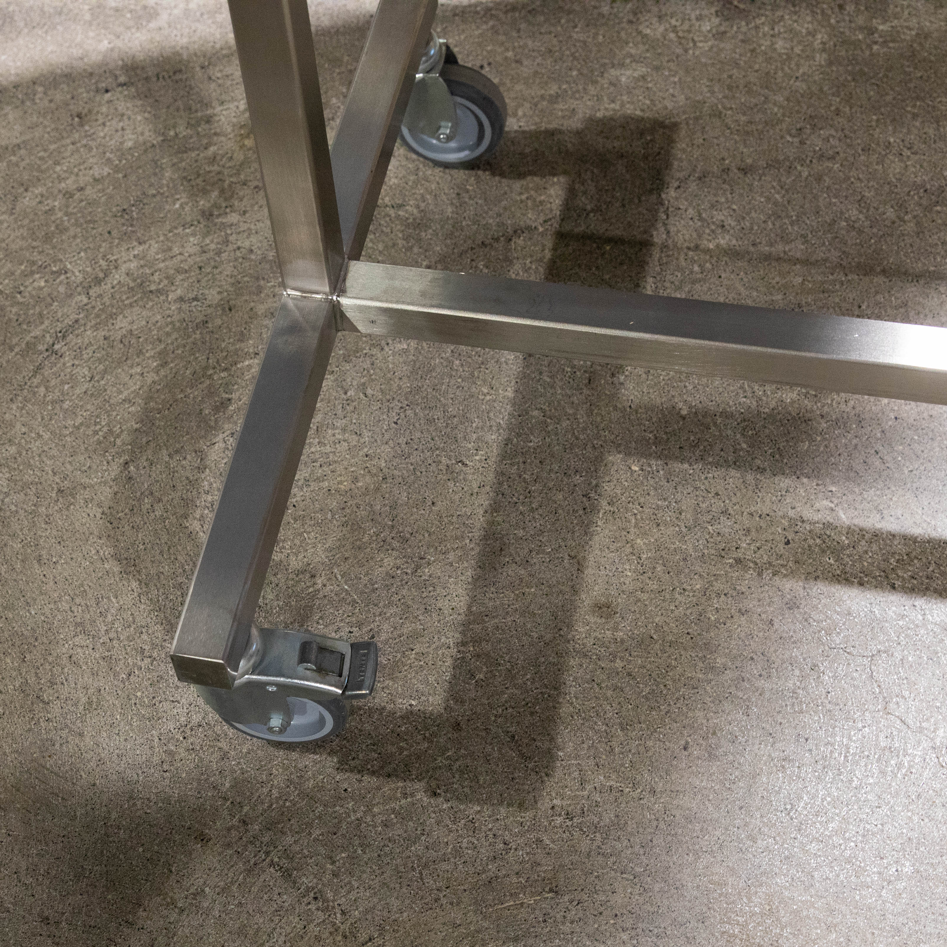 Nahaufnahme des Fußbereichs eines Kleiderständers aus glänzendem Chromstahl auf grauem Betonboden. Quadratische Metallrohre bilden eine stabile Ecke mit grauer Lenkrolle samt Arretierung. Die Rolle ist fixiert, eine weitere teilweise sichtbar.