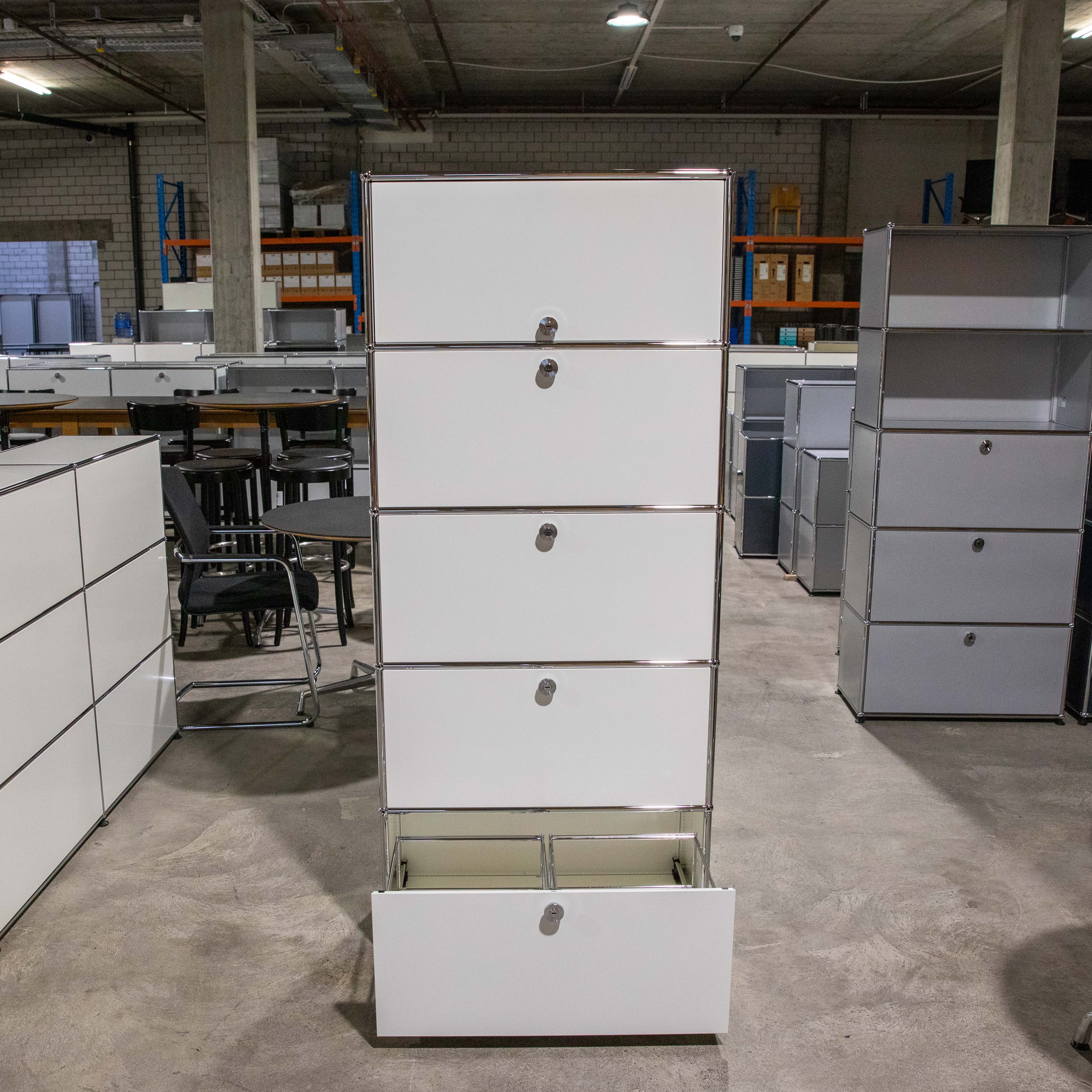 Hohes, weißes USM Haller Regal mit fünf Fächern aus weißen Metallplatten und Chromrohren. Unten offene Ausziehtür mit Hängeregistratur. Oben vier Schranktüren mit silbernen Schlössern. Auf Betonboden.
