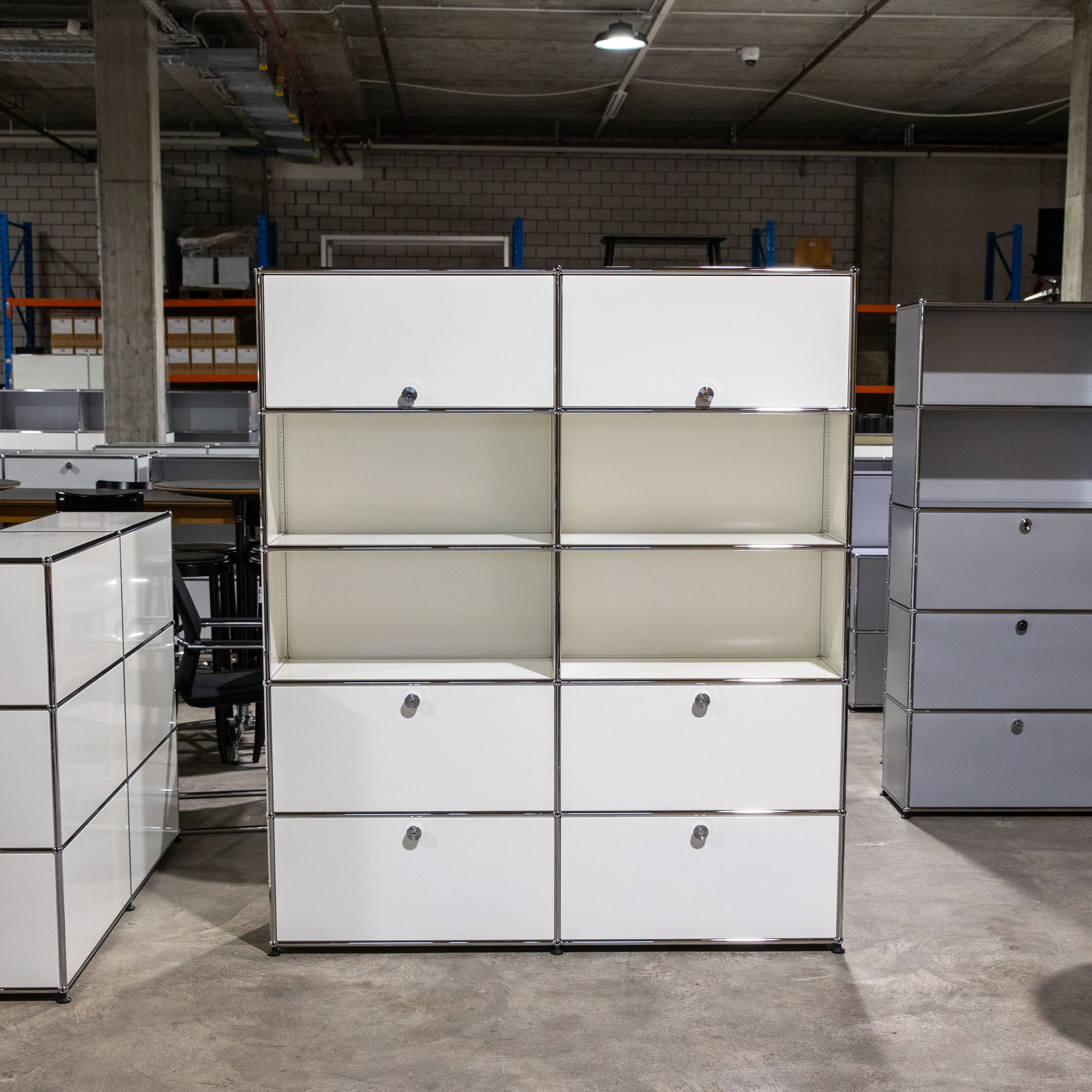 Großes weißes USM Haller Regal mit 10 Fächern, modular in 2 Spalten und 5 Reihen. Oben und unten mit weißen Türen/Schubladen und silbernen Griffen/Schlössern, mittig 2 offene Fächer. Silberner Rahmen, auf Betonboden.