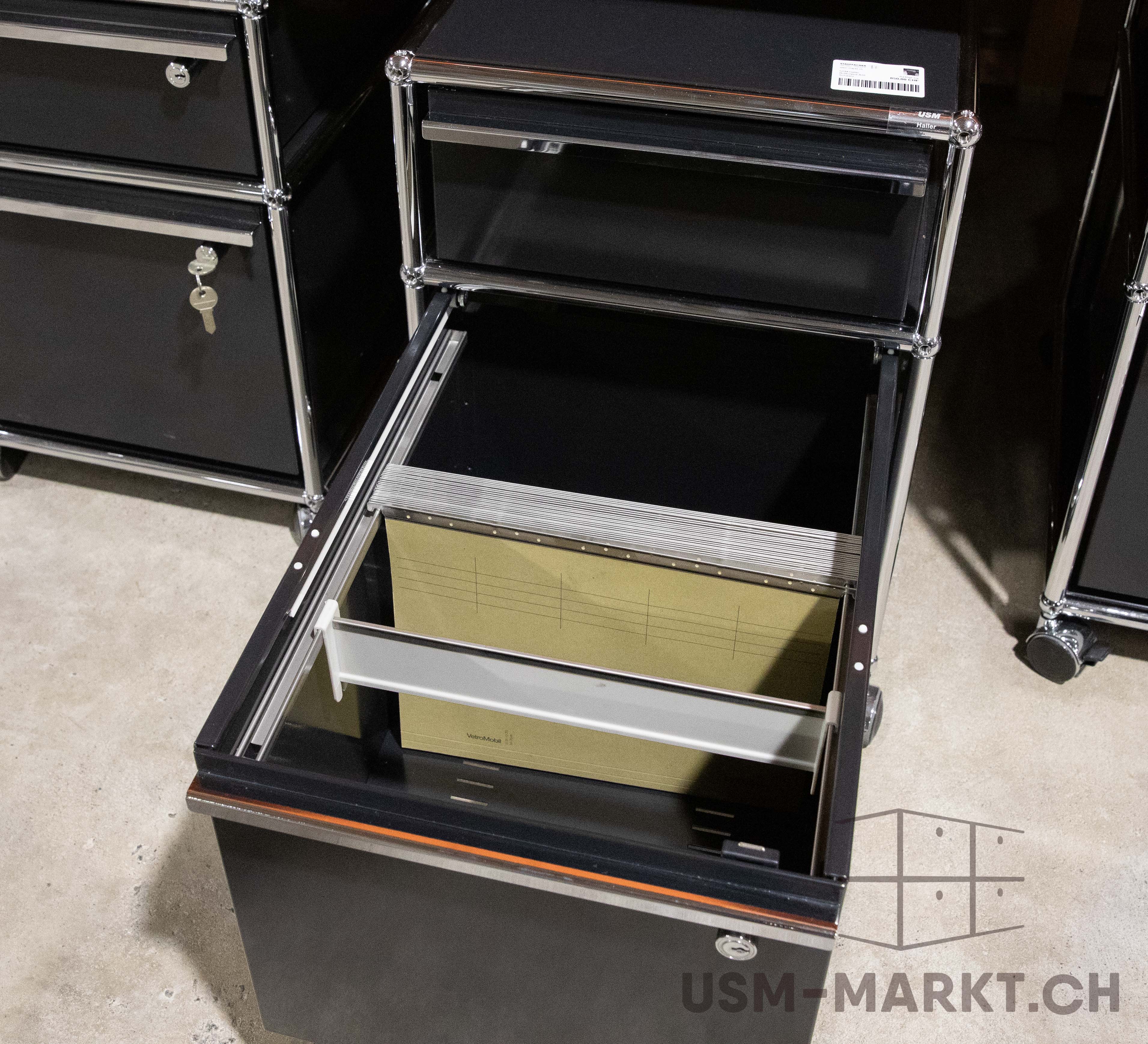 Schwarzer USM Haller Rollcontainer auf hellem Boden, Schublade geöffnet mit beigen Hängemappen. Modulares Möbel mit verchromten Streben und Rollen. Links ein geschlossener Schrankteil mit Schlüssel im Schloss.
