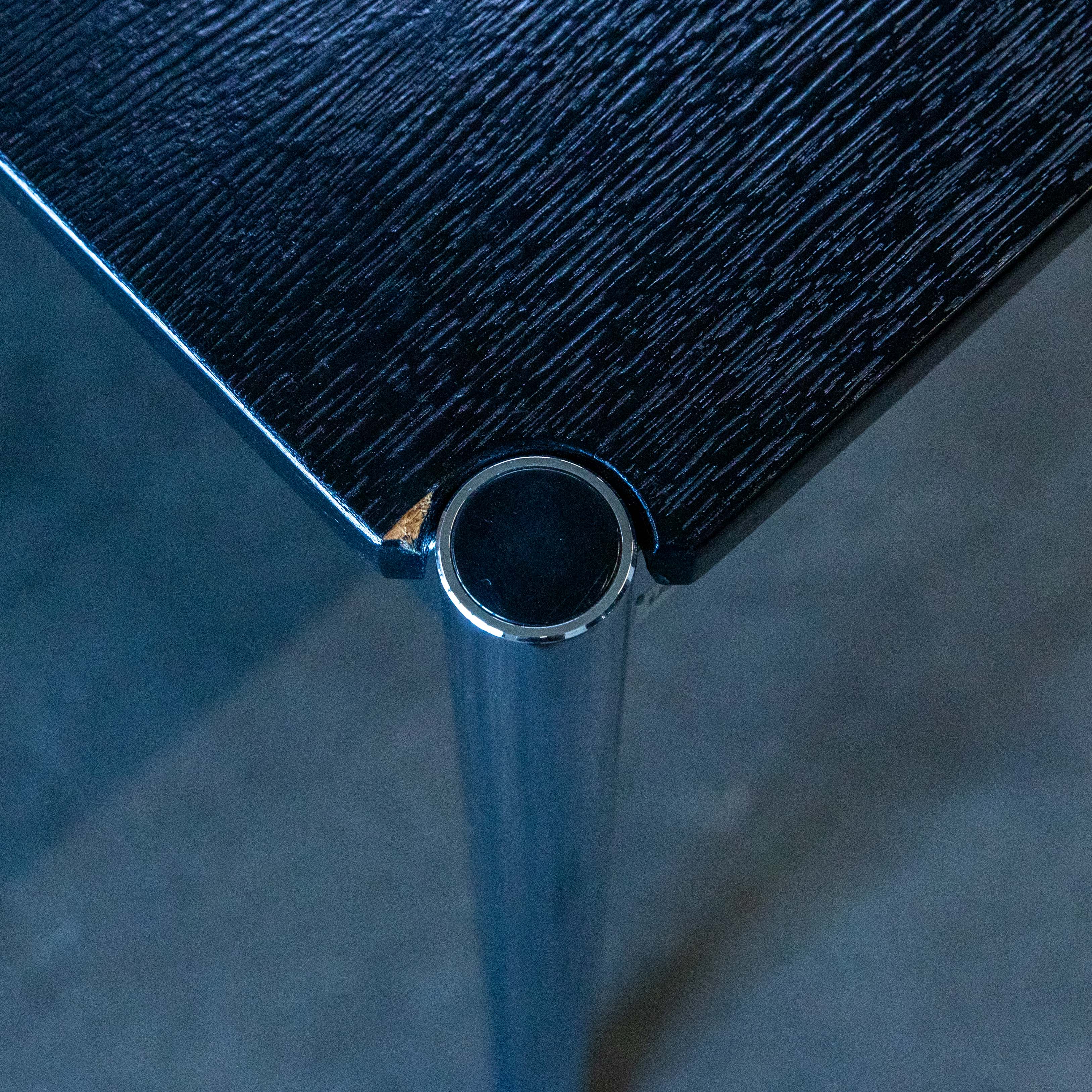 Gros plan sur un coin d'une table USM Haller noire. Le plateau en chêne laqué noir laisse apparaître le grain du bois. Un pied chromé brillant est relié au plateau par un élément métallique rond. Petite éraflure sur la laque.