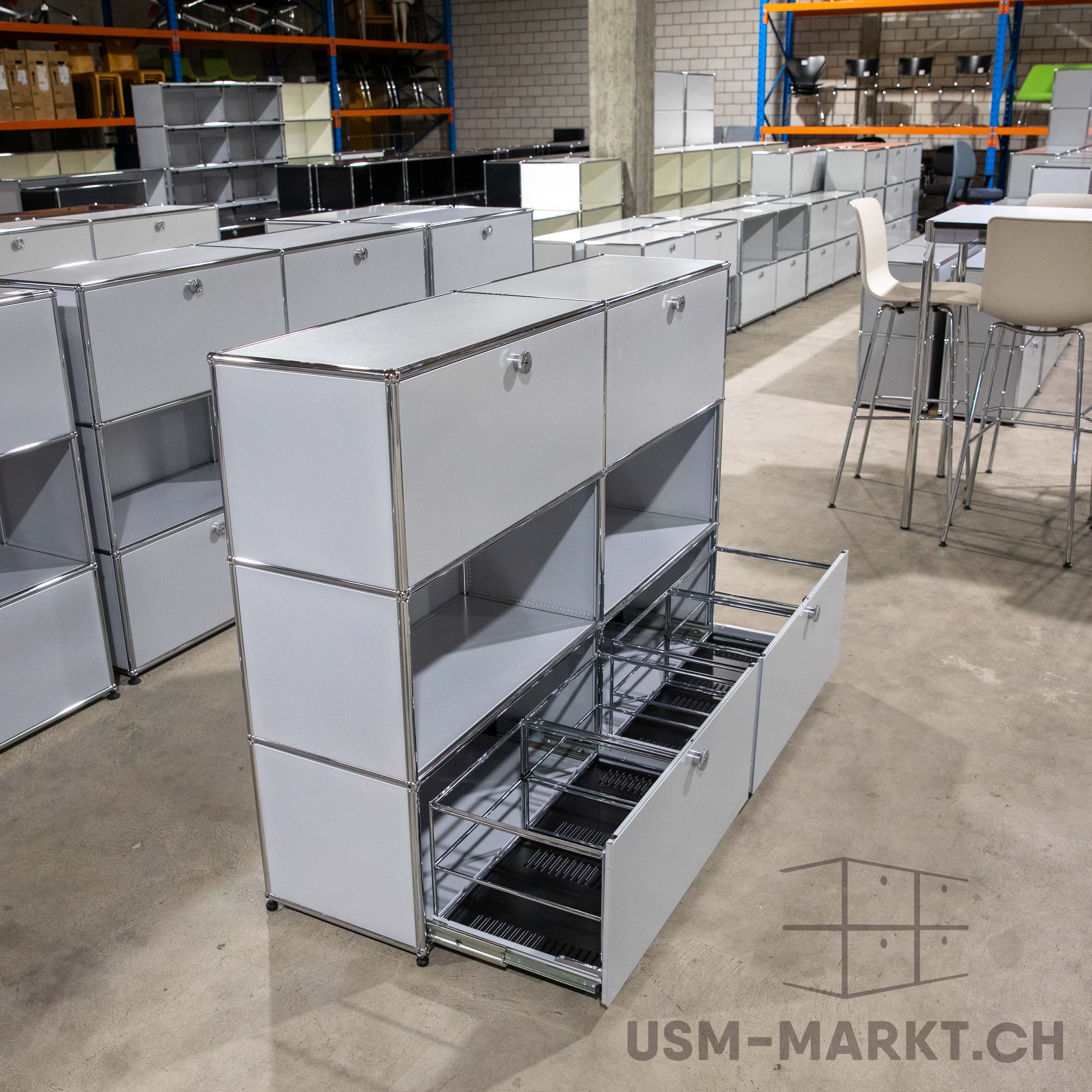 Modernes modulares Schranksystem in Weiß mit glänzenden Chromrahmen in einer Lagerhalle. Mehrere Fächer, teils mit Türen, teils offen, unten eine halb ausgezogene Schublade mit Metallschienen für Hängeregister.