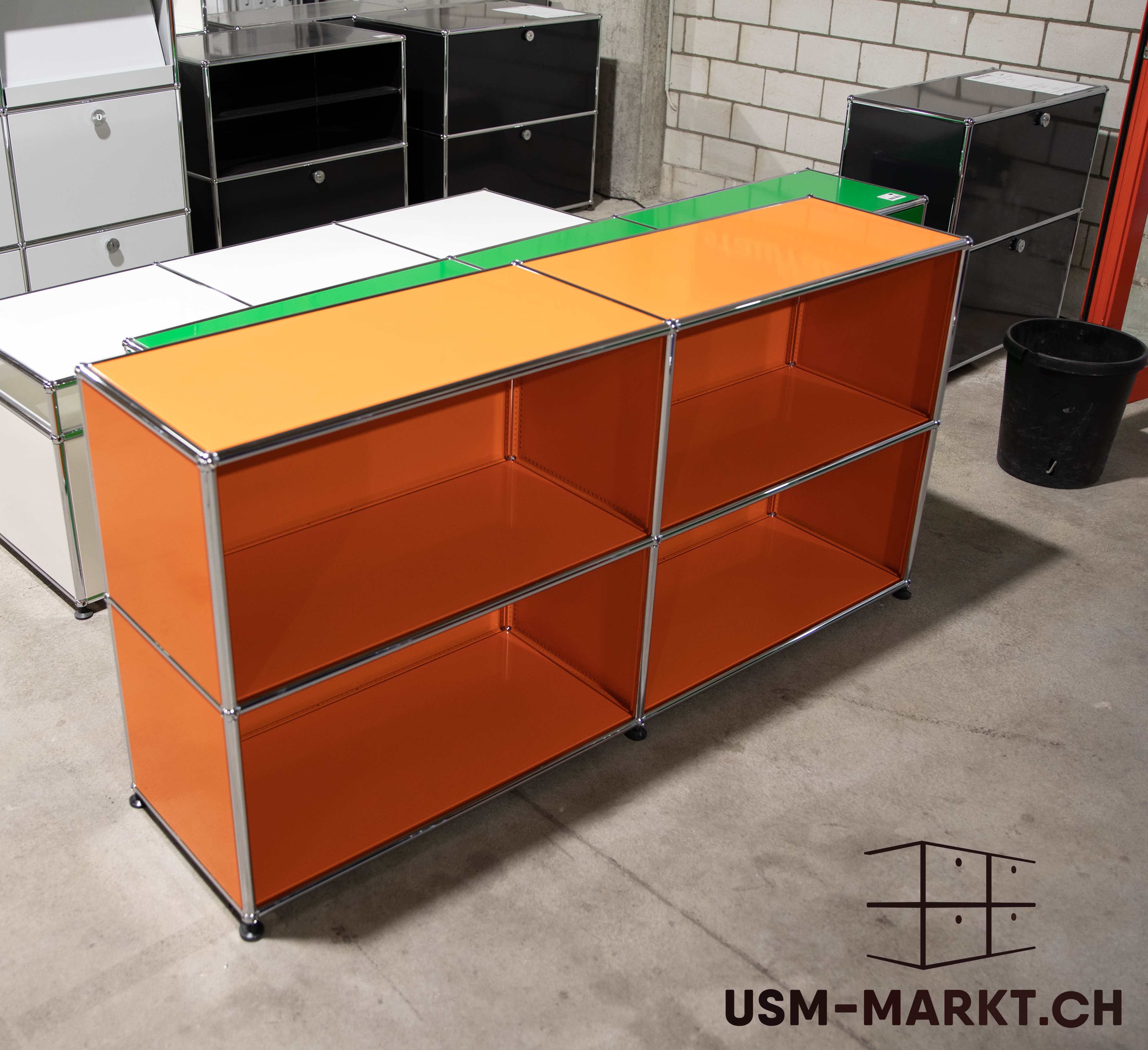 A long, modular USM Haller shelving unit in bright orange and green, two-tiered with open compartments and chrome-plated steel tubes, on a concrete floor. In the background, more USM Haller furniture in white and black against a white brick wall.