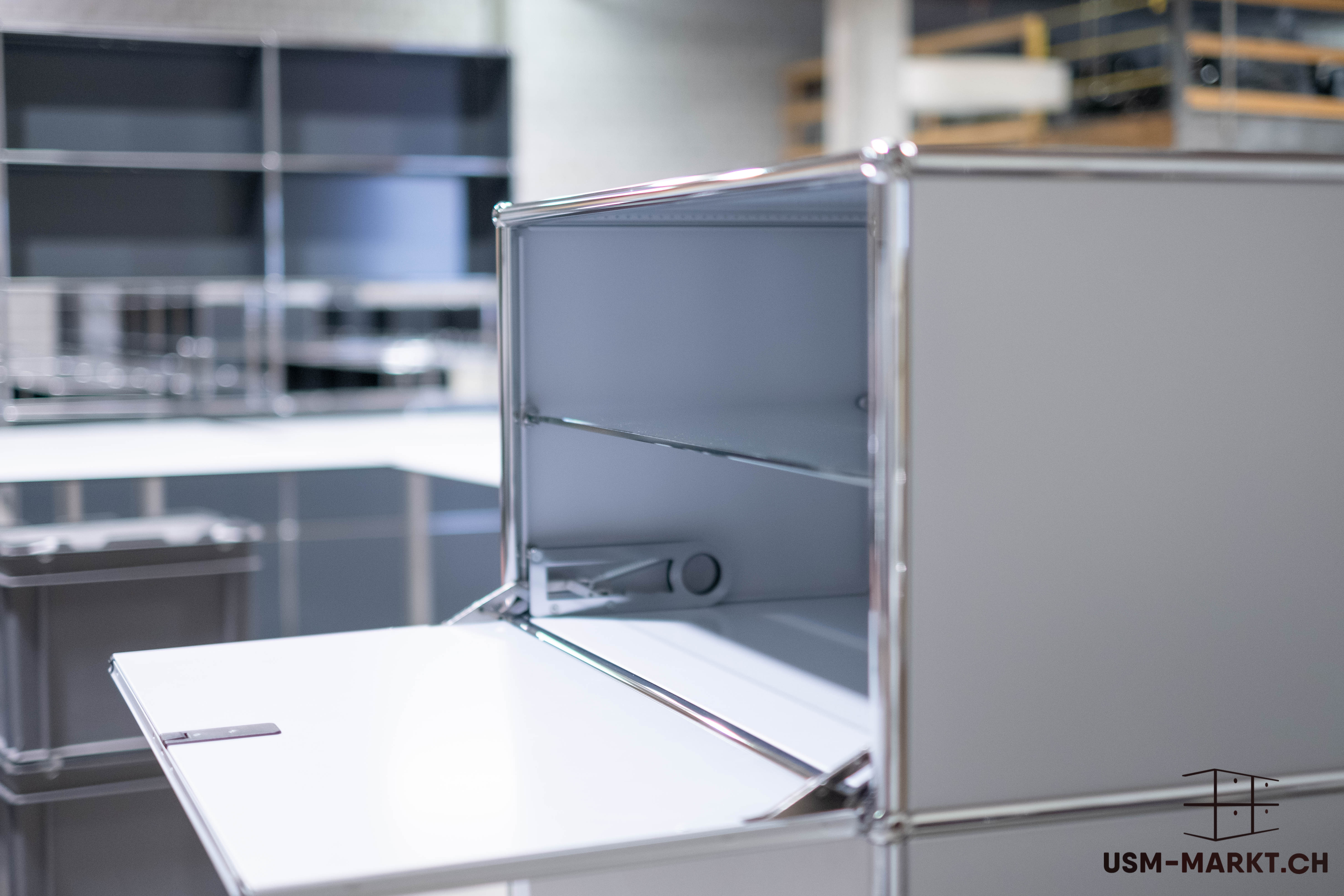 A light-coloured, modular USM Haller unit with an open hinged door, revealing an empty compartment with a shelf. The chrome-plated connecting elements and matt finish are clearly visible. Further shelves can be seen in the blurred background.