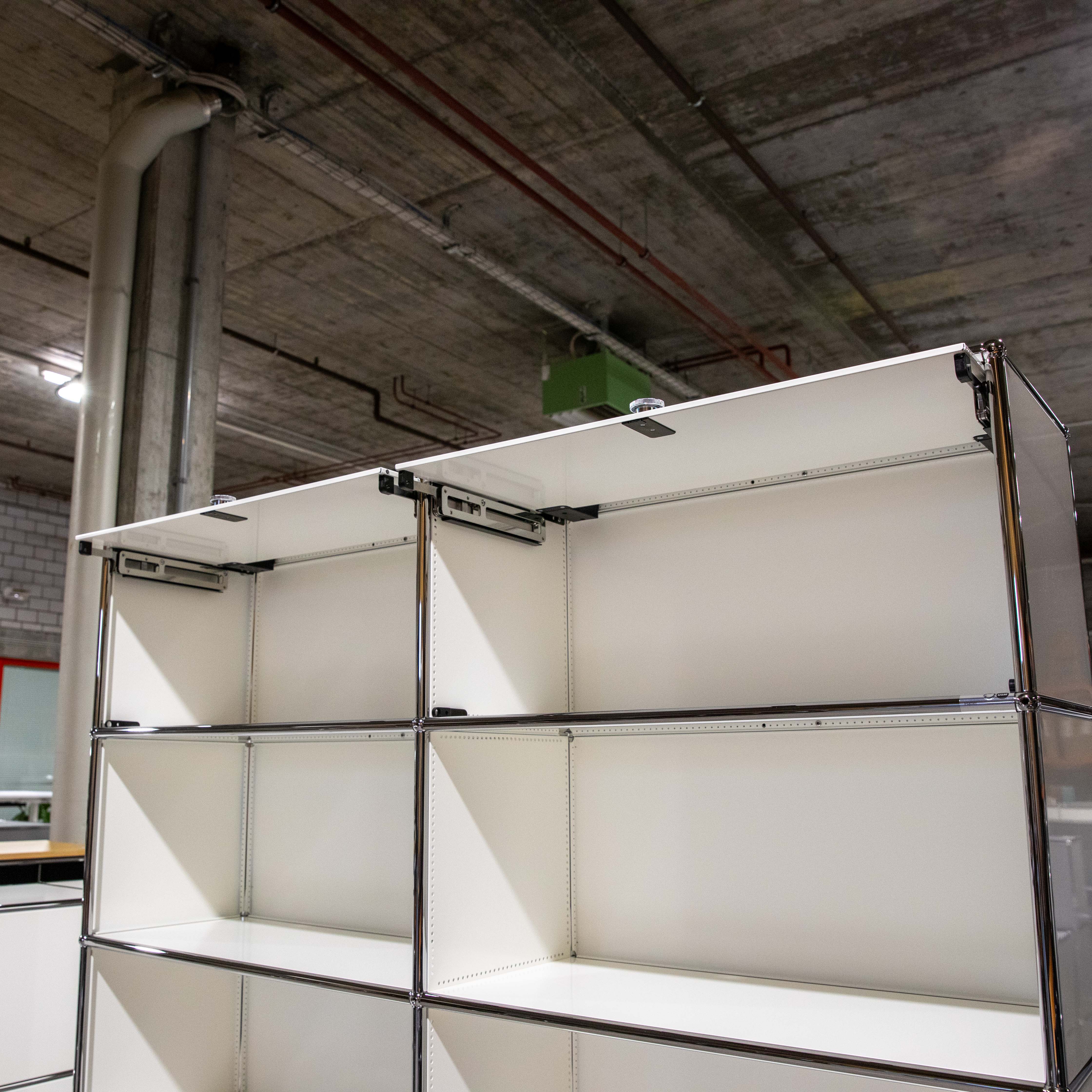 Detailansicht eines weißen USM Haller Regals in Top-Occasion Zustand. Modulares Möbel mit offenen Fächern und glänzenden Chromstangen als Rahmen. Oben sind Mechaniken für Auszüge und Klapptüren erkennbar.