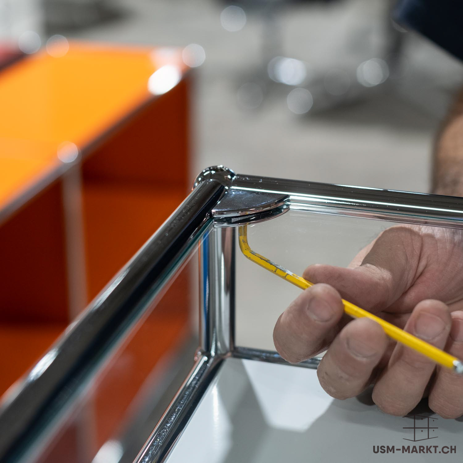 Una mano assembla un mobile e inserisce una chiave a brugola gialla a forma di L in un giunto cromato lucido. Il giunto collega i tubi cromati lucidati di un sistema modulare. Sullo sfondo, un pannello arancione.