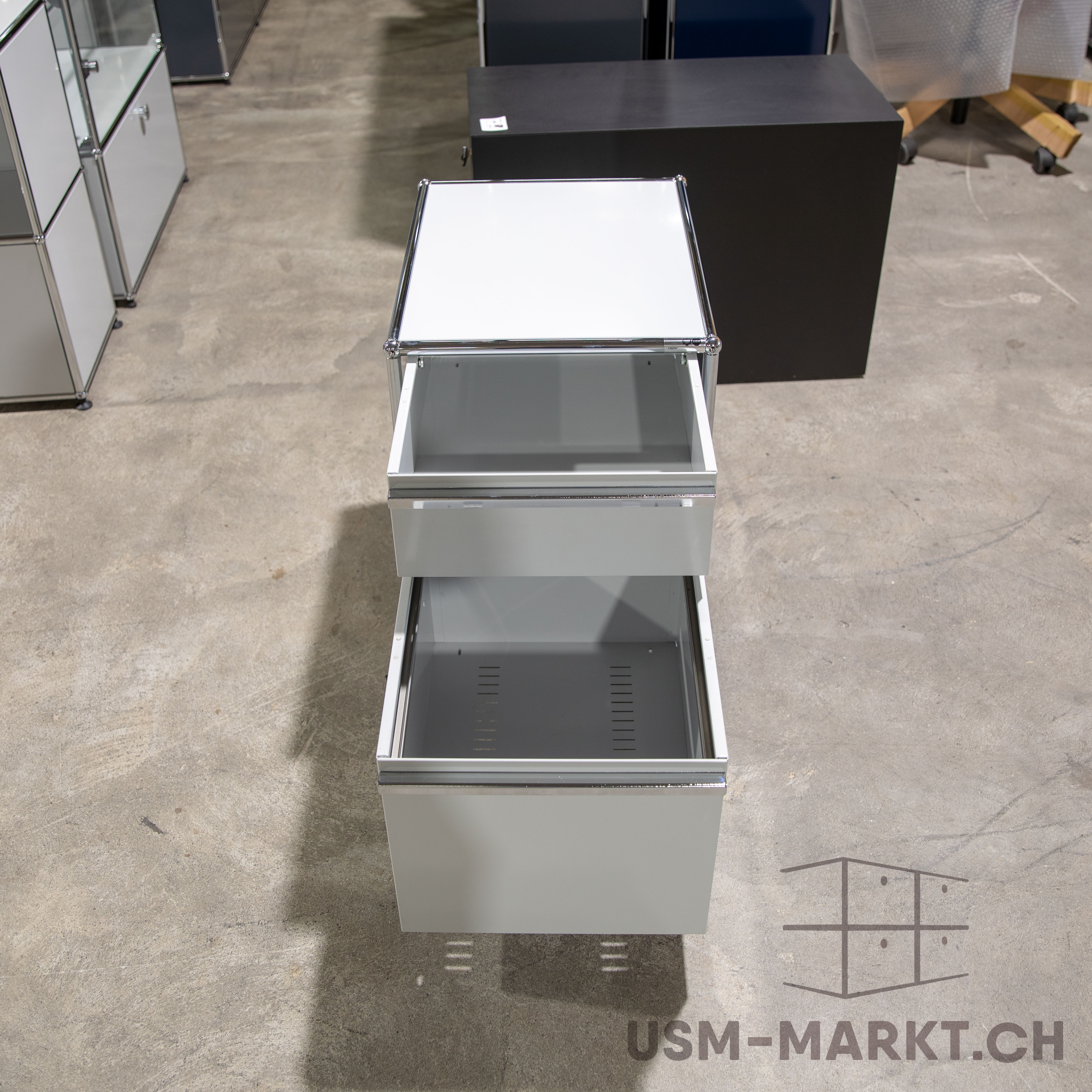 Hellgrauer, moderner Rollcontainer mit zwei offenen Schubladen auf grauem Betonboden. Oben teils herausgezogen, unten ganz geöffnet, beide leer. Verchromte Griffe und Rahmen, minimalistisch, Hintergrund unscharf.