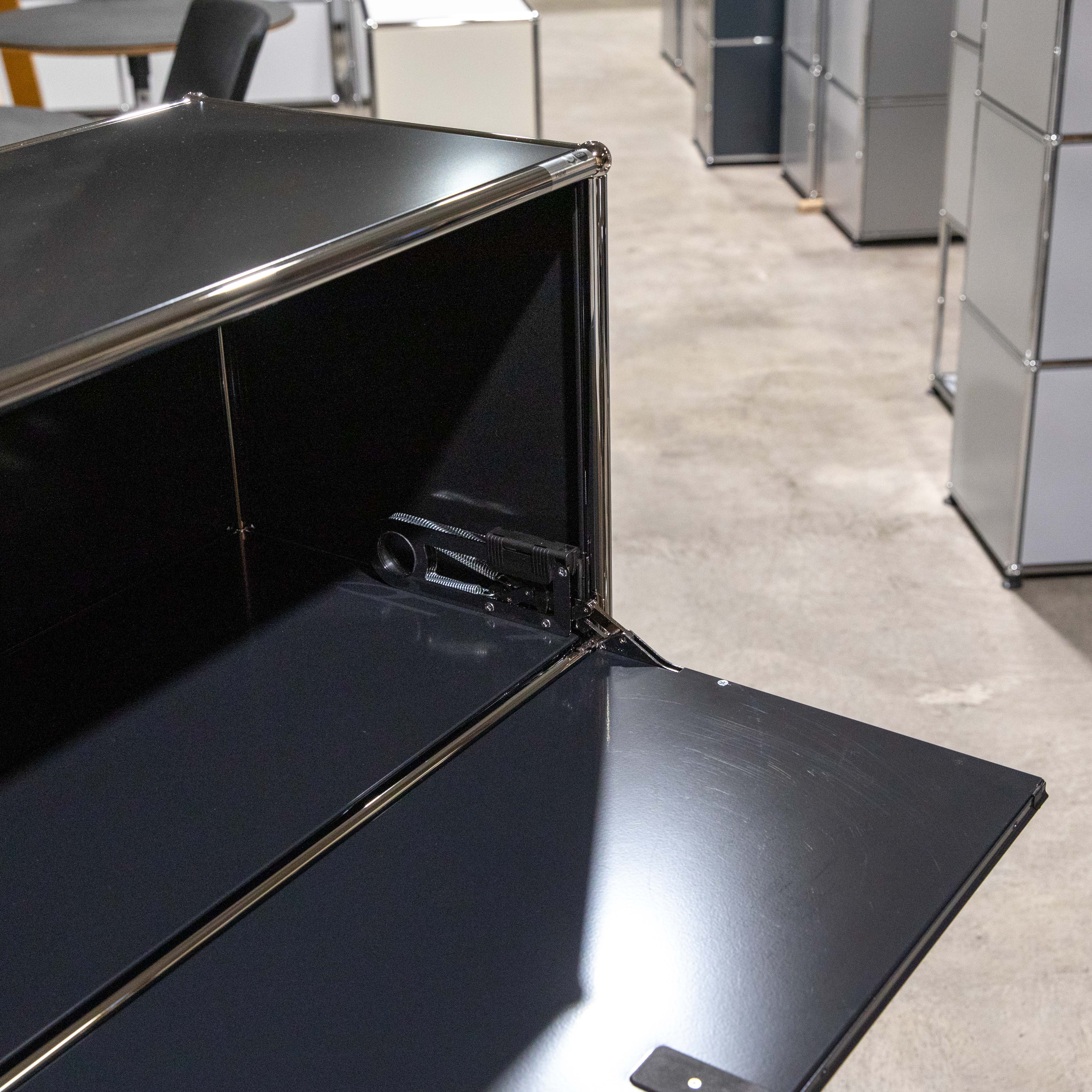 Nahaufnahme eines geöffneten, schwarzen USM Haller Regalmoduls. Klapptür nach unten geöffnet, dunkles Fach sichtbar. Chromscharniere und SilentClose-Mechanismus mit Federn detailliert. Matte Oberfläche mit Glanzreflexen.