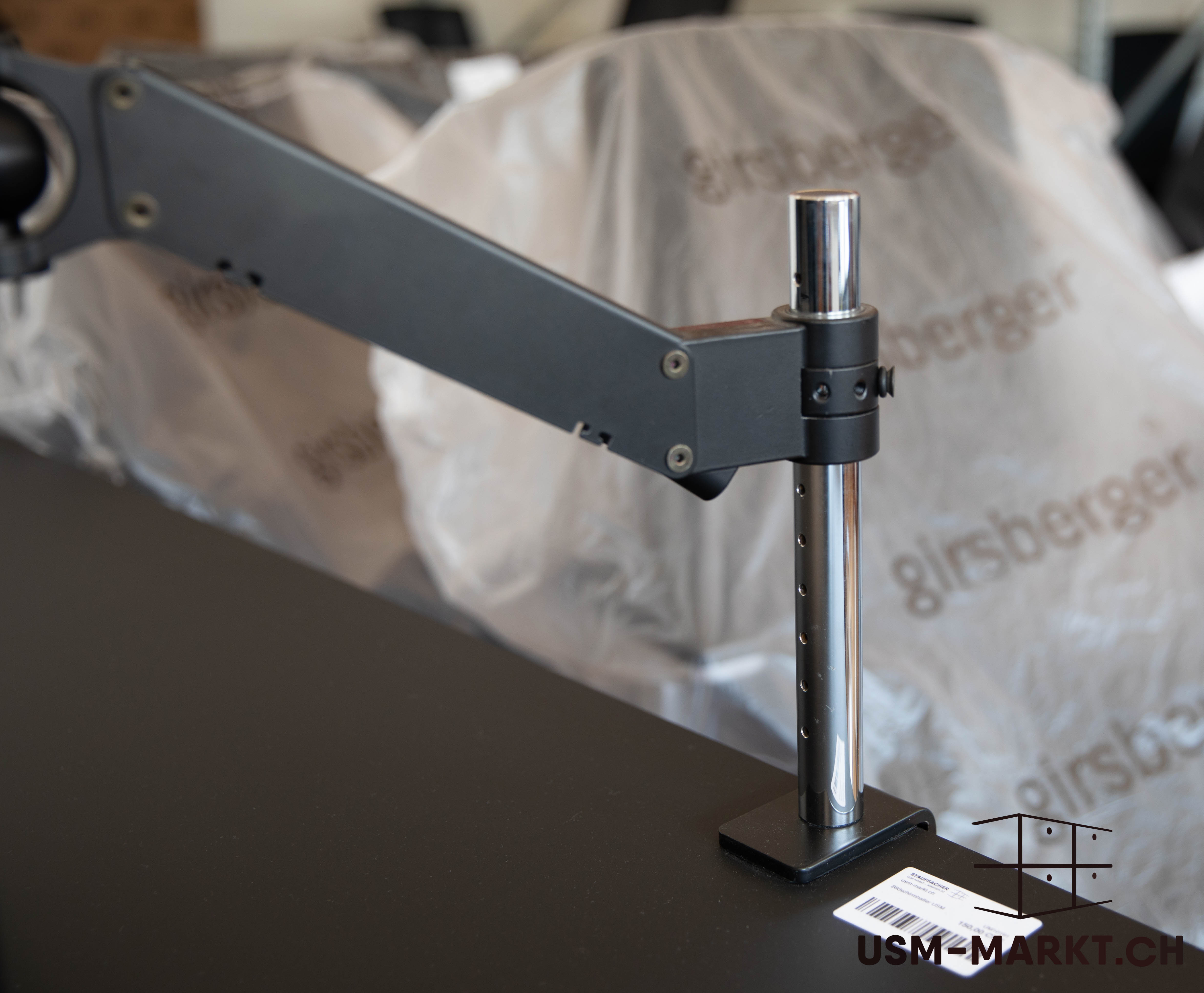 Un bras de moniteur noir et réglable est monté sur une surface sombre, avec un montant en chrome brillant et un bras articulé noir mat. À l'arrière-plan, flou, on aperçoit des objets « Girsberger » recouverts d'un film plastique.
