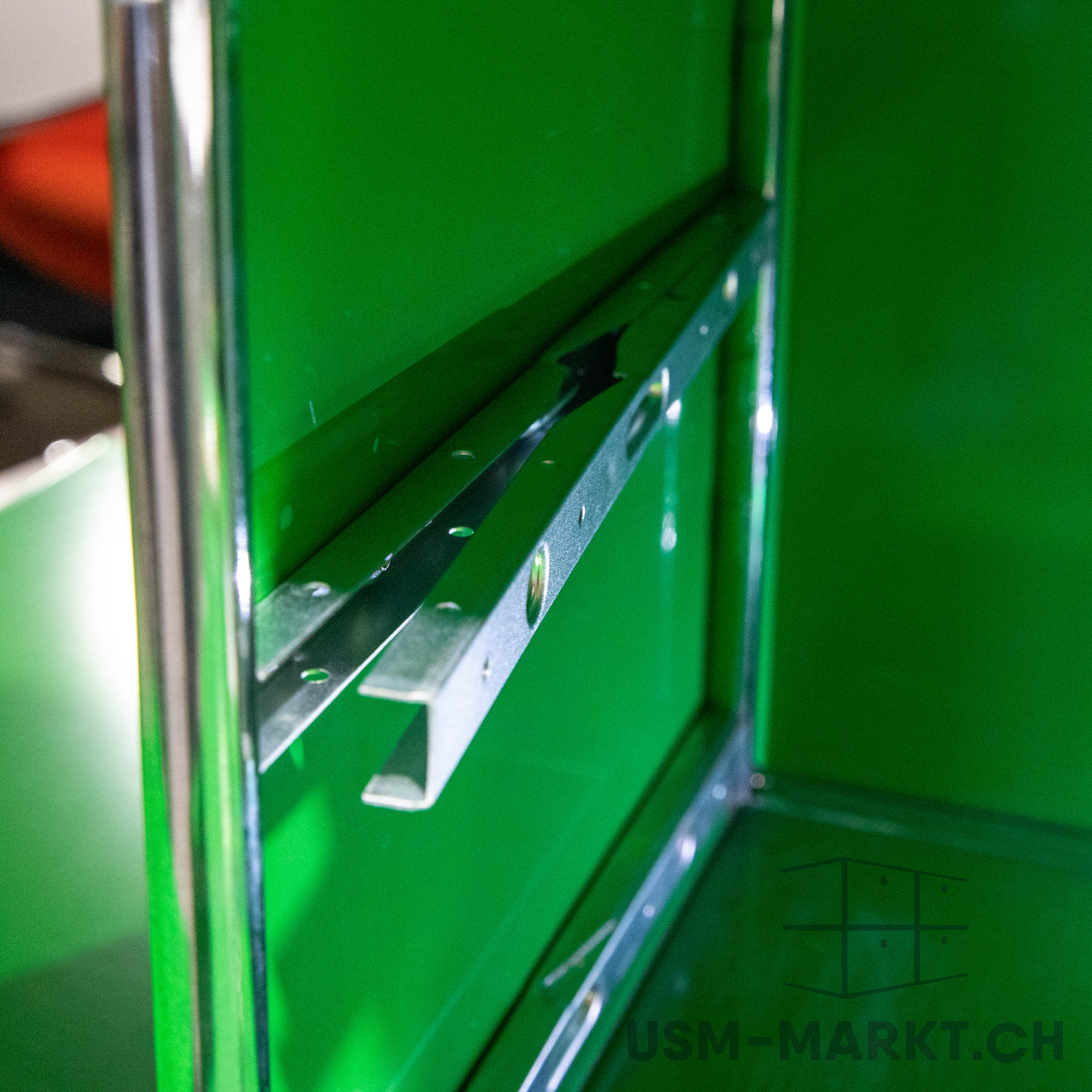 A detailed view of a bright green modular unit with an open interior. Two horizontal, perforated metal rails for adjustable positioning of shelves are visible. On the left is a polished chrome rail.