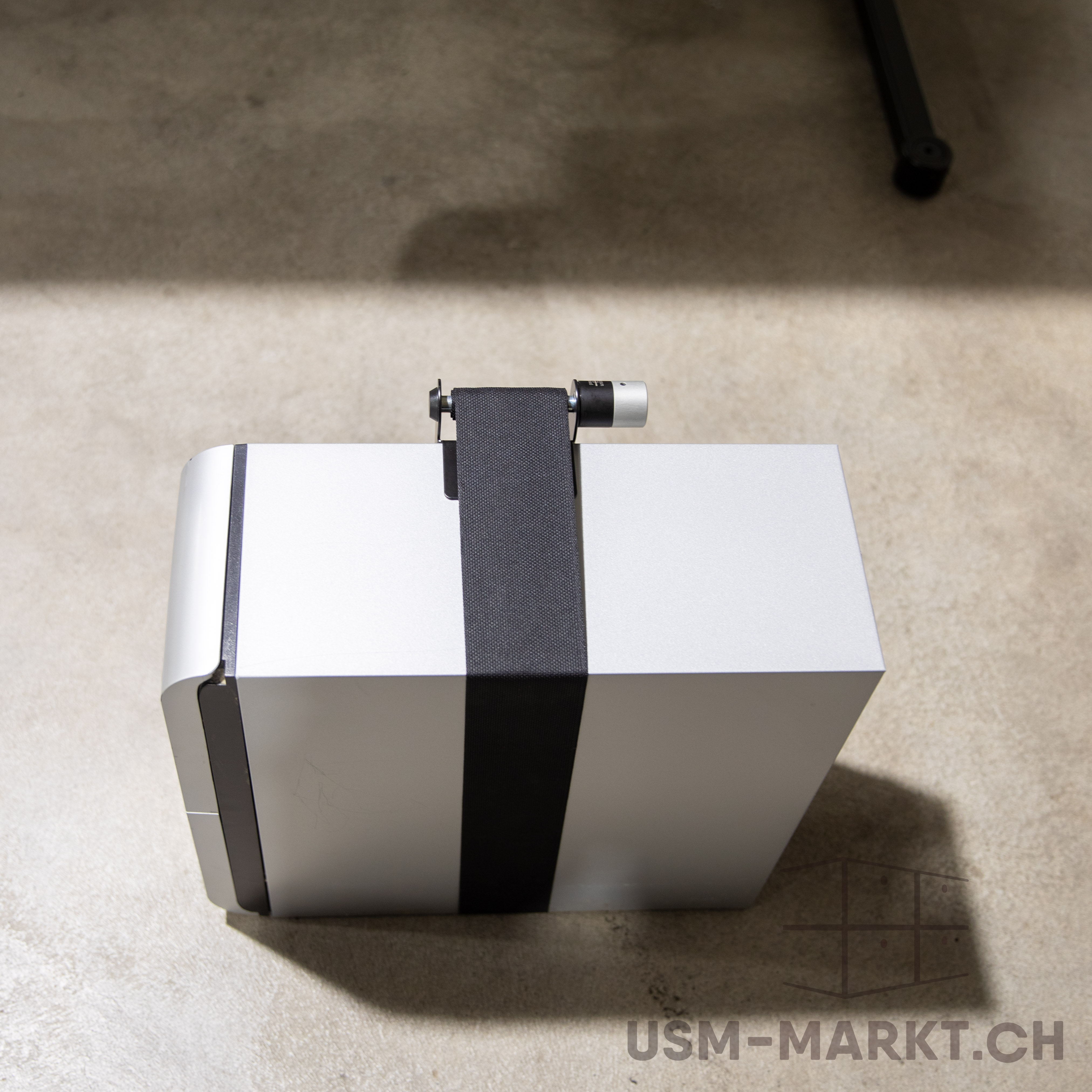 Ein rechteckiger, hellgrauer Behälter liegt auf hellbraunem, strukturiertem Boden. Ein schwarzes Stoffband ist vertikal um den Behälter gespannt und oben von einem silbernen-schwarzen Klemmmechanismus gehalten.