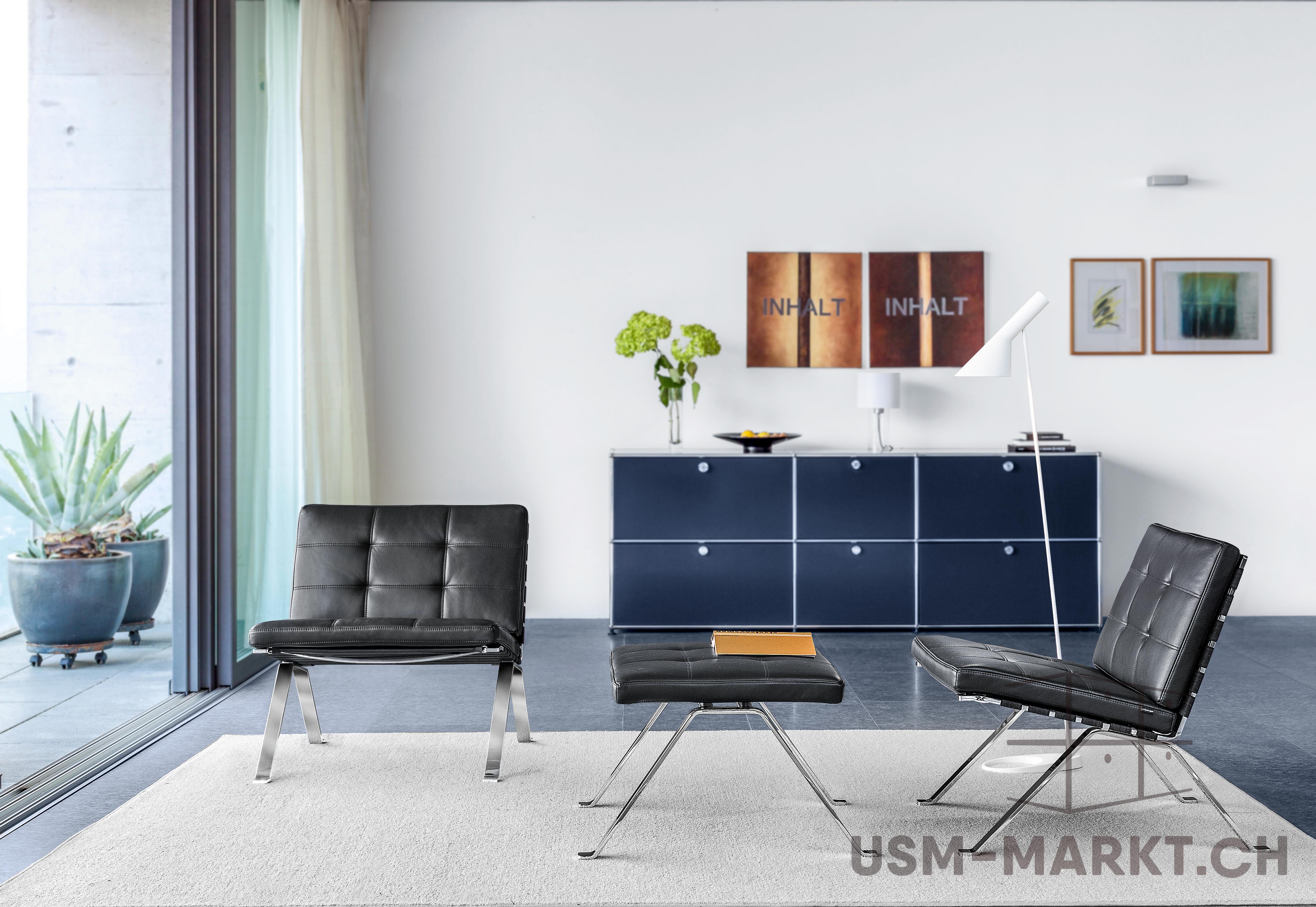 Due poltrone Barcelona in pelle nera con struttura cromata e pouf abbinato su tappeto grigio chiaro in una stanza moderna. Sullo sfondo, credenza USM Haller blu scuro con oggetti e opere d'arte. A sinistra, porte scorrevoli in vetro sul balcone.