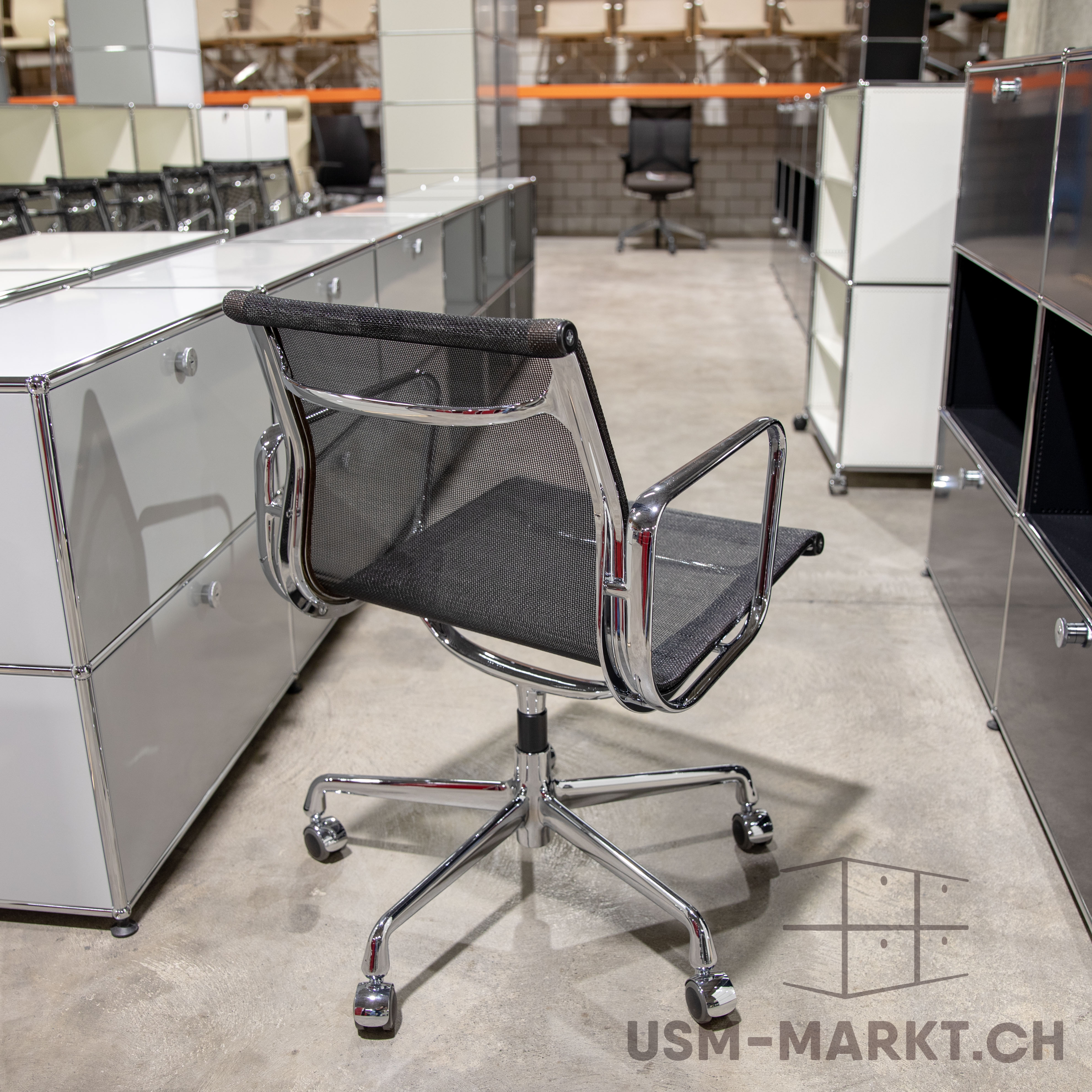 Sedia da ufficio moderna in rete nera e struttura cromata al centro di un pavimento in cemento grigio, con base su rotelle. Sullo sfondo, mobili modulari USM Haller bianchi, grigi e neri come scaffali e cassettiere.